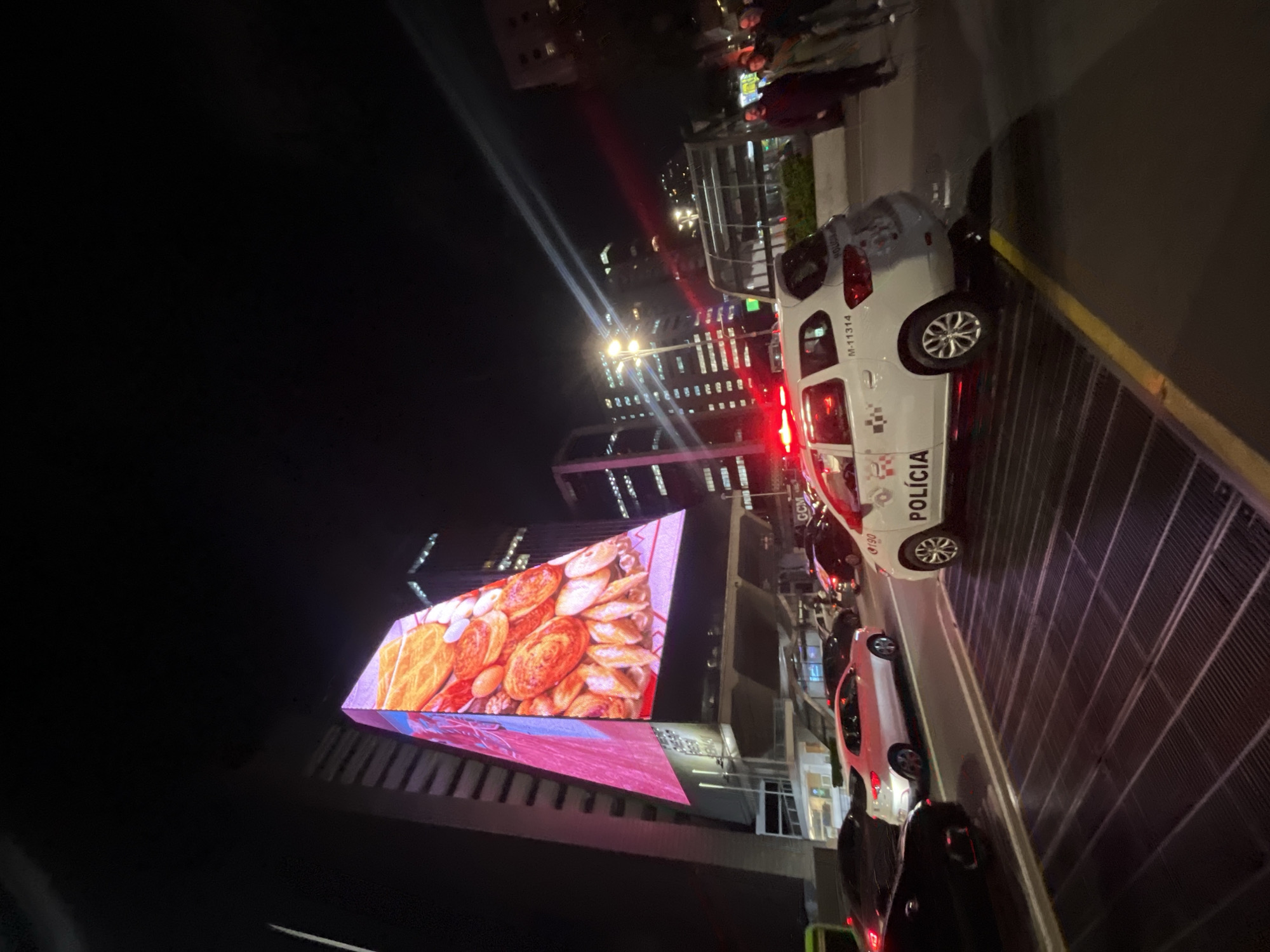 At night, the image depicts a city street with illuminated buildings in the background. A large electronic billboard displays an image of pastries. Several vehicles, including a white car marked "POLÍCIA", are parked along the curb. People stand near the building, near a metal barrier. The setting appears to be urban.