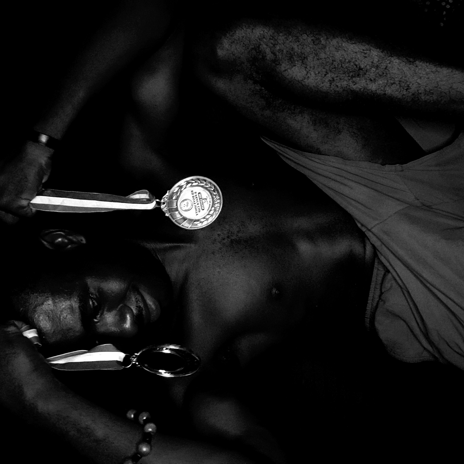 The image is a black-and-white photograph of a dark-skinned person lying down, presumably male, with their head to the left. The person's face is visible, showing a closed eye and relaxed expression. Two medals are positioned on the person's chest and near their head. One medal rests directly on the person's upper chest, while the other appears to be held up near the person's face. The person wears a wristband on their left wrist and a beaded bracelet on their right wrist. The person's upper body is bare, with a garment draped across the lower portion of their body. The setting is dark and obscured, making it difficult to determine the environment. There is no clear indication of Narayi, Nigeria.