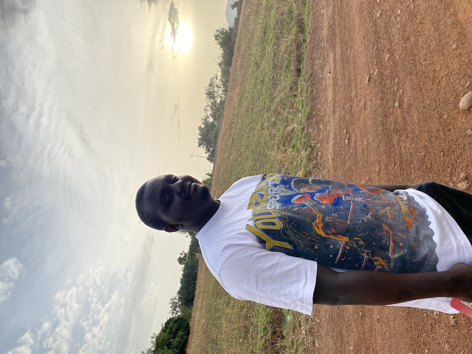 A person is standing in a rural area near Bukuru, Nigeria. The individual is wearing a white T-shirt with a large graphic print. The landscape includes dirt roads, grasslands, sparse vegetation, and trees in the background. The sky is overcast with a visible sun.