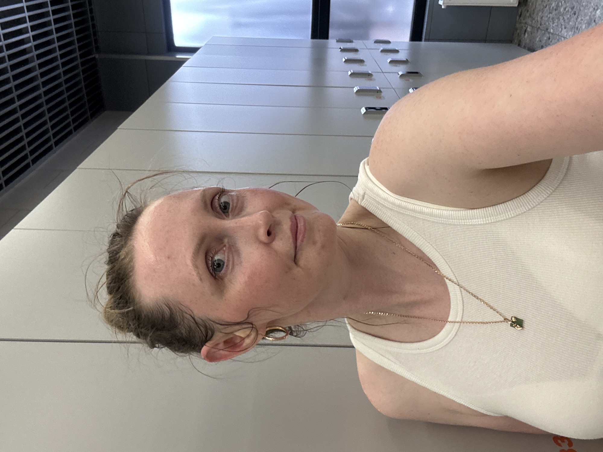 A woman with light skin and reddish-blonde hair pulled back in a bun is captured from the shoulders up, facing the camera at an angle. Her hair is slightly messy, with wisps framing her face. She wears a white tank top and a delicate gold necklace with a square green pendant. Her right ear is adorned with a gold hoop earring. The background consists of a row of grey lockers, characteristic of a gym or changing room. The ceiling above is a dark, gridded structure. The overall impression is that of a candid moment captured in a public facility. The image does not provide any specific details or context related to the city of Votkinsk, Russia.
