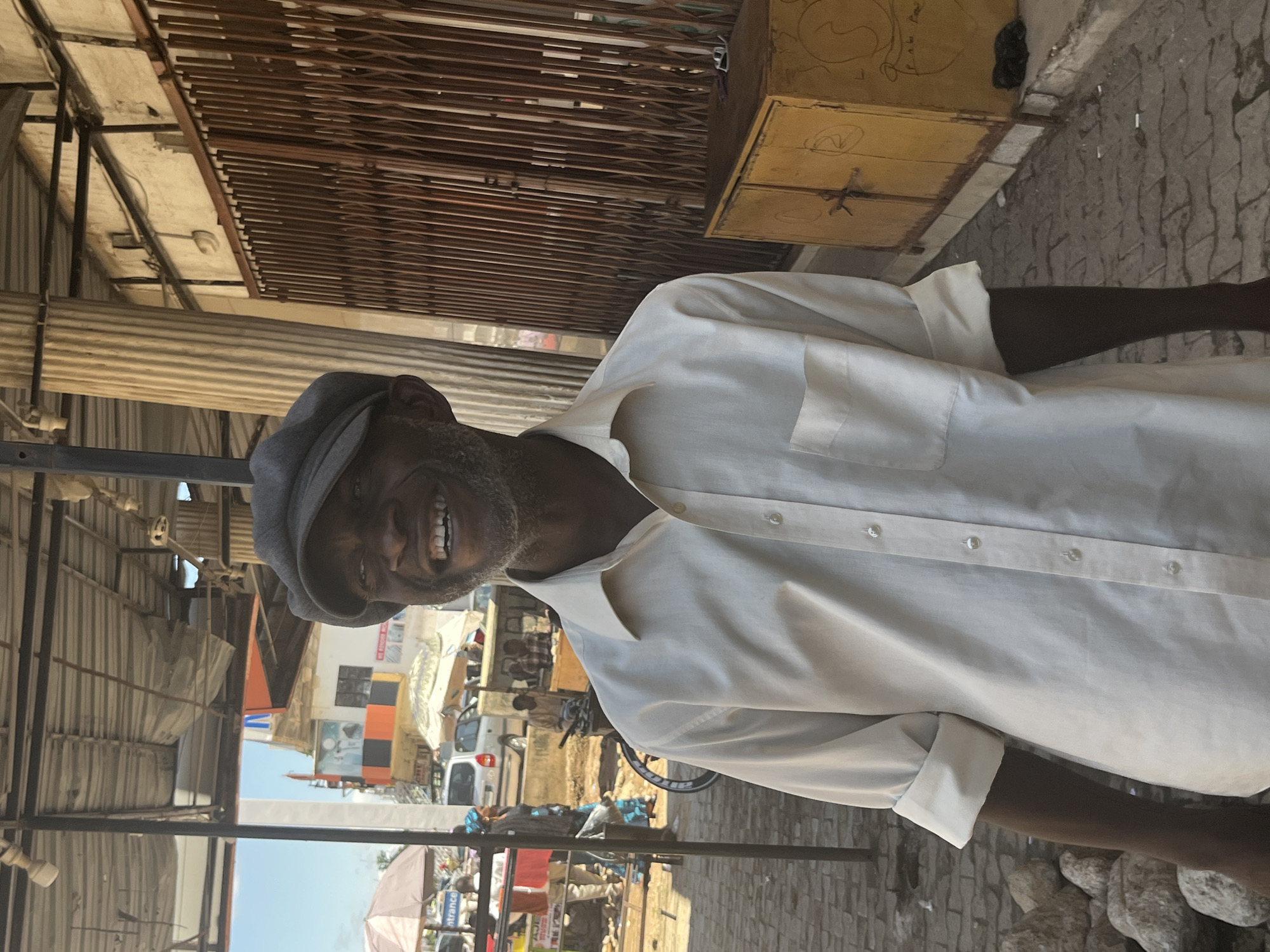 The image shows a Black man wearing a light-colored button-down shirt and a dark flat cap, smiling and looking towards the viewer. He has a beard and graying stubble. The background appears to be an outdoor market or street scene, likely in Jos, Nigeria, given the context of other images from this source. To the left, there are stalls with umbrellas and vehicles, including a white car and a bicycle. The man is standing on a paved surface, with rocks to his left. To the right, there is a tall corrugated metal structure and a yellow metal cabinet with writing on it. The overall lighting suggests it is daytime.