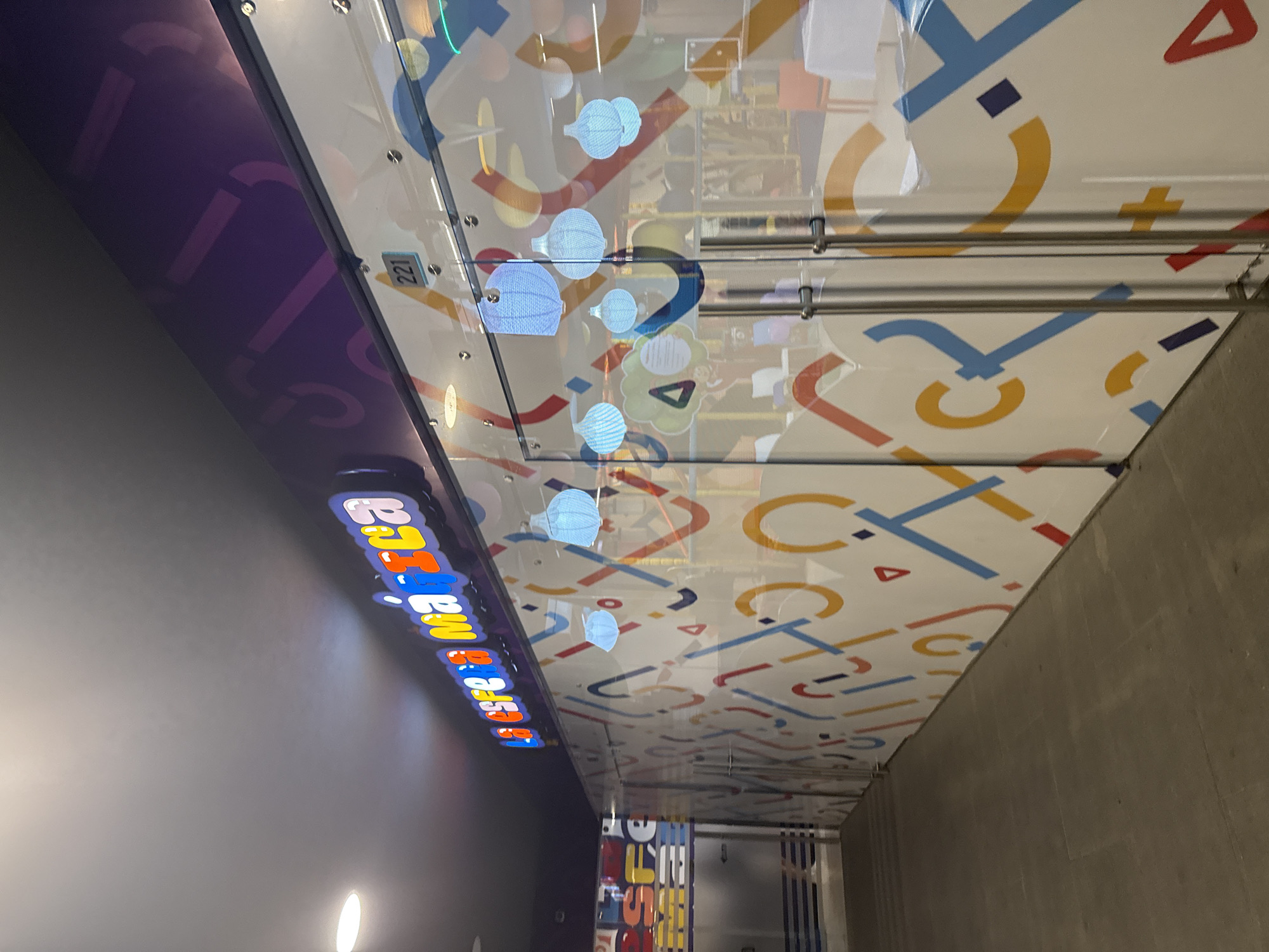The image depicts an indoor commercial corridor in San Bernardino Tlaxcalancingo, Mexico, leading towards a storefront. The primary subject is a brightly lit storefront named "LA ESFERA MÁGICA," displayed on a prominent sign above the entrance in multi-colored, stylized letters. The storefront features double clear glass doors with vertical brushed metal handles, and the unit number "221" is affixed above the right door. Both the glass facade and the adjacent wall to the left are covered with a repeating graphic pattern of abstract shapes, including arcs, lines, triangles, and squares, rendered in blue, red, yellow, orange, and white on a white background. Through the clear glass, the interior of the establishment is visible, featuring blurred furniture elements and multiple light-blue, spherical, lantern-like decorative objects suspended from the ceiling. The corridor floor consists of large, dark gray square tiles. The overhead ceiling is dark, with a circular recessed light fixture visible and illuminated in the upper left portion of the frame. No individuals are present in the scene.