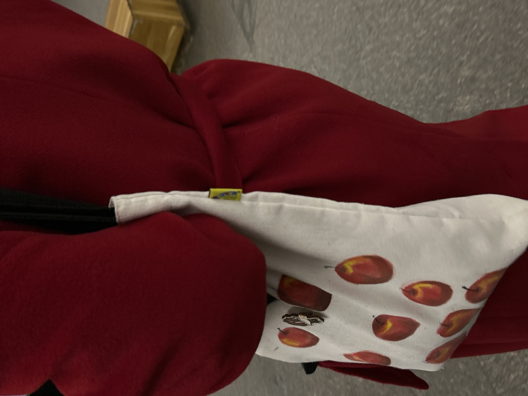The image provides a close-up, angled view of a person's attire and accessories, set in what is contextualized as Saint Petersburg, Russia.
The primary subject is a rich, dark red fabric, likely a coat or jacket, which fills a significant portion of the frame. This fabric appears soft, possibly wool or fleece. Intersecting with the red fabric is a band of white fabric, which is patterned with numerous stylized fruits. These fruits are painted in shades of red, orange, and yellow, resembling apples or pears, each with a small dark stem or end point. This white fabric could be part of a tote bag, a scarf, or an accent on the clothing.
A small yellow tag with blue markings is visible where the red and white fabrics meet, suggesting a brand label, though the text on it is illegible. Further down on the white fabric, a small metallic pin or brooch is attached. It appears silver-toned and depicts a small, intricate shape, possibly a stylized animal or symbol, but its exact form is difficult to discern due to its size and angle.
The background is largely blurred and greyish, possibly a wall or floor. A wooden object, partially visible in the upper left background, is also out of focus. No specific activity is taking place; the image is a static shot of clothing details. The time of day and weather are not discernible from this indoor close-up.
