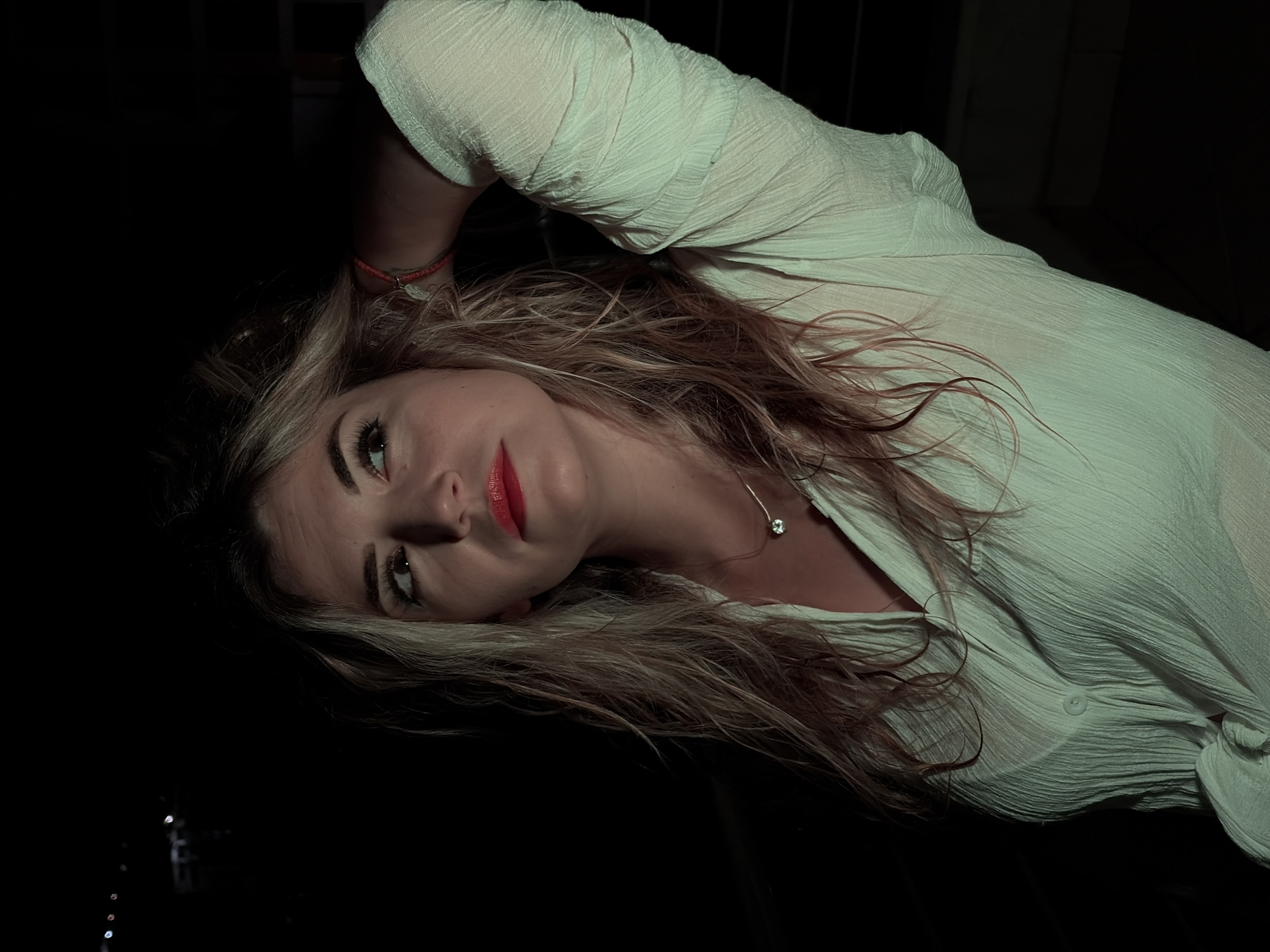 The image captures a woman at night in Budva, Montenegro. She has long, wavy hair, blonde and light brown in tone, appearing somewhat tousled. Her face, illuminated from the left, features red lipstick and visible eye makeup. Her gaze is directed upward and to her right. She wears a white, textured, long-sleeved shirt, which appears semi-sheer, revealing a lighter garment underneath. Around her neck is a delicate silver-toned necklace with a single, clear, round pendant. A red string bracelet with a small charm adorns her right wrist. Her right arm is bent, with her hand placed behind her head. The background is predominantly dark, with a few faint, out-of-focus lights visible in the upper left, indicative of a distant urban or coastal environment at night.