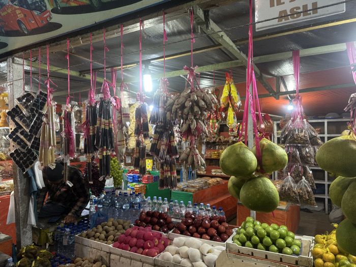 A bustling market stall displays an array of goods. Packaged incense sticks and dried goods hang from the ceiling with pink ties. Fruit is neatly arranged below, including apples, lemons, and pomelos. Bottled water sits atop the fruit. A vendor in a plaid shirt and a hat is behind the stall. A sign is visible on the ceiling.