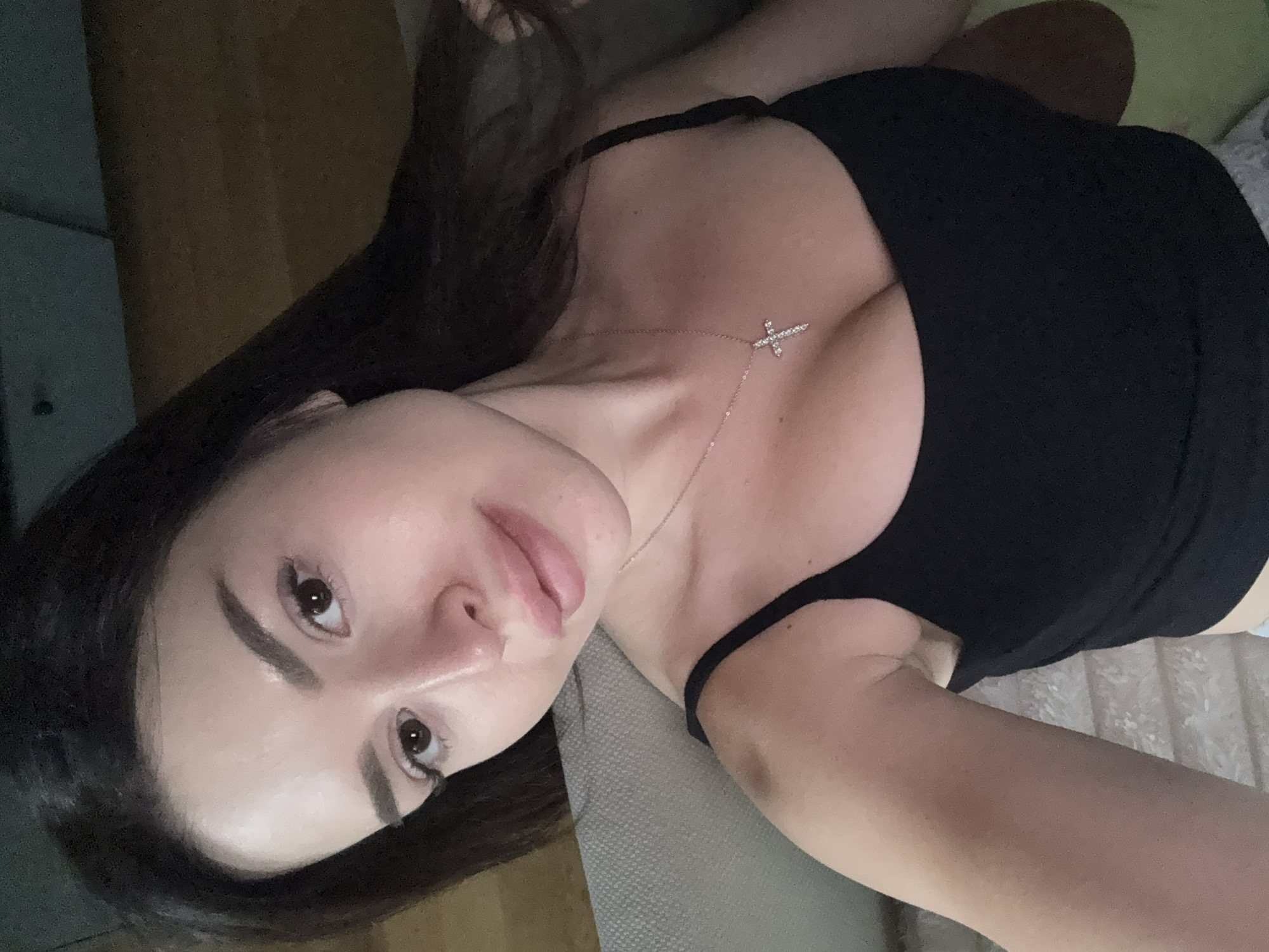 A young woman with dark hair and light skin is taking a selfie indoors. She is wearing a black tank top and a diamond cross necklace. The background is blurred but appears to be a living room with a couch. There are no visible signs of the city of Prague.