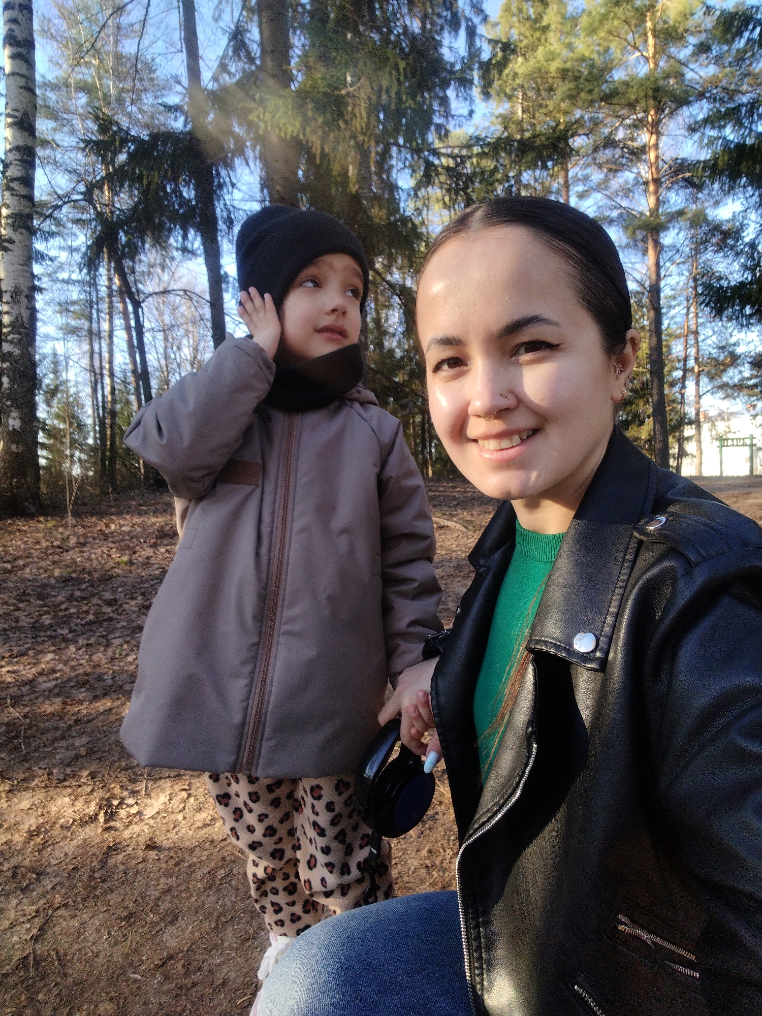 A selfie photograph shows an adult woman and a child outdoors. The woman is on the right side of the image, wearing a black leather jacket over a green sweater and blue jeans. The child, on the left, wears a grey jacket, a black beanie, and animal-print pants. The child's right hand is raised to their ear. The woman is holding headphones. Behind them is a wooded area with bare trees and foliage on the ground. The image was taken in Noviy, Russia.