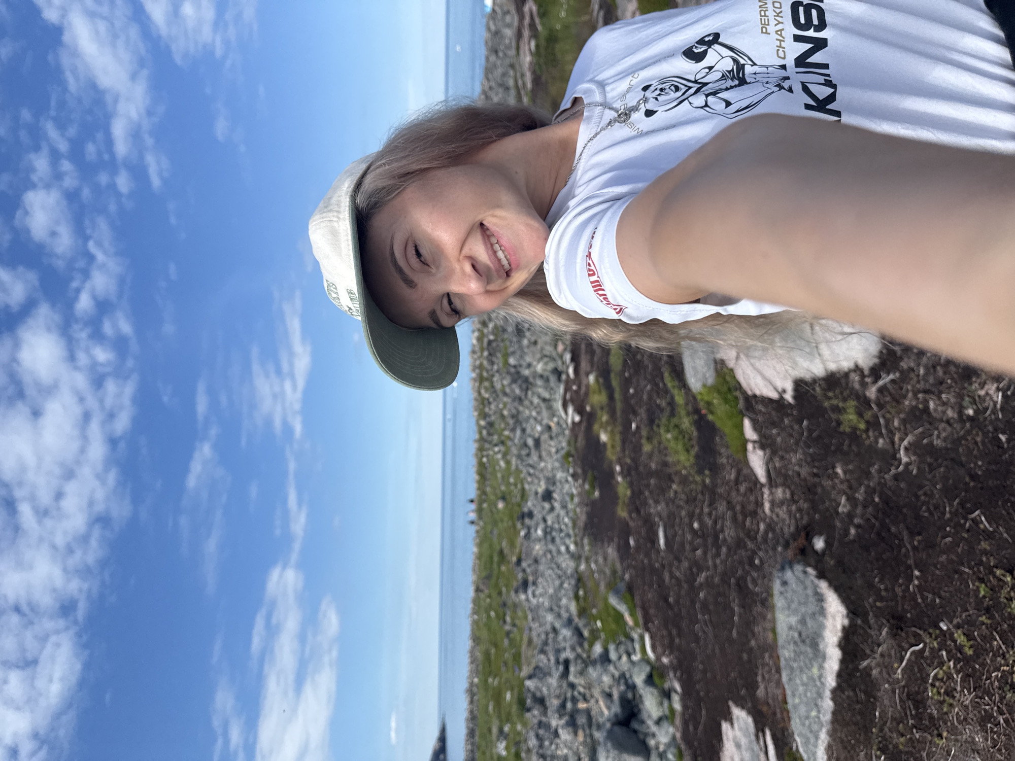 A blonde woman wearing a white t-shirt and a light-colored baseball cap takes a selfie on a rocky, coastal landscape under a blue sky with scattered clouds. The woman is smiling and looking at the camera. Her t-shirt has a graphic of a bear in boxing gloves and text that reads "PERM CHAYKO KINS". In the background, the ocean is visible, with a rocky shore leading to the water. Some distant figures are standing by the water's edge. The scene appears to be in Teriberka, Russia, a coastal town known for its rugged natural beauty.