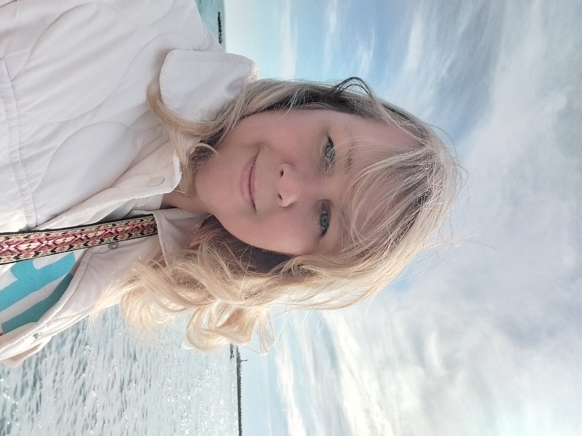 The image is a close-up selfie of a fair-skinned woman with long, wavy blonde hair and blue-green eyes, captured outdoors. She has a gentle, pleasant smile. The woman is wearing a light-colored, possibly white or cream, quilted jacket, underneath which a light blue and white patterned top is visible. A decorative strap with a geometric pattern in shades of red, pink, black, and cream crosses her chest.
The background features a large body of water, which, given the context, is likely the Black Sea in Sochi, Russia. Above the water, a bright blue sky is visible with scattered white, wispy clouds. The lighting suggests it is daytime. The overall mood conveyed by the woman's expression is one of contentment or calm. There is no visible text in the image. The image is presented rotated 90 degrees clockwise.