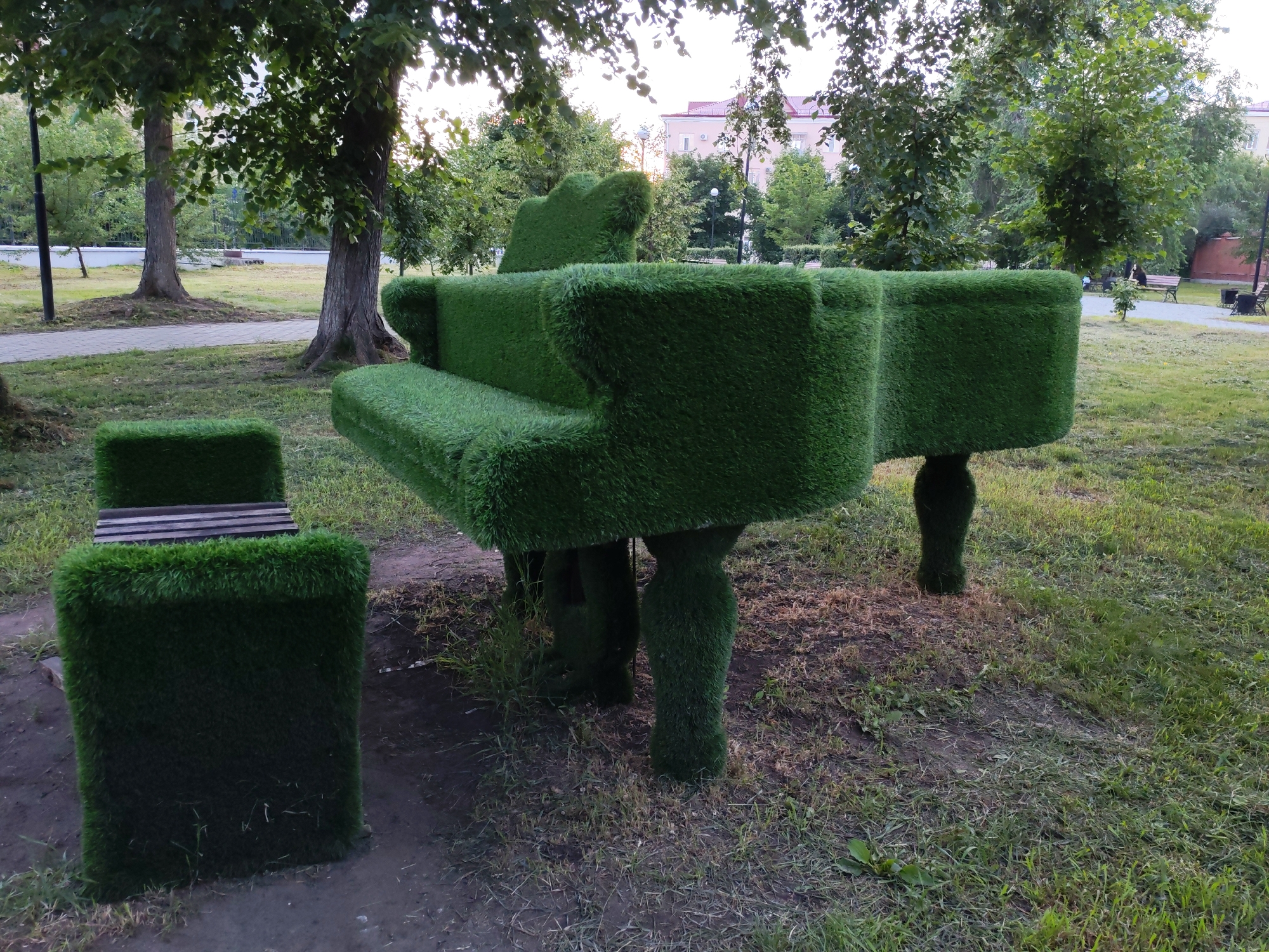 The image shows a whimsical, life-sized piano sculpture covered in artificial grass, placed outdoors in a park-like setting. The piano is a grand piano model, complete with a bench that is also covered in artificial grass. The sculpture is situated on a patch of dirt and grass, with trees and foliage surrounding it. In the background, there is a paved walkway, more trees, and a building with a pink roof. The lighting suggests it is daytime, possibly late afternoon, with soft sunlight filtering through the leaves of the trees. There is no visible text in the image, and no people are present, giving the scene a serene and artistic atmosphere. The unusual material choice for the piano sculpture suggests it is an art installation or a creative decorative element in the park.