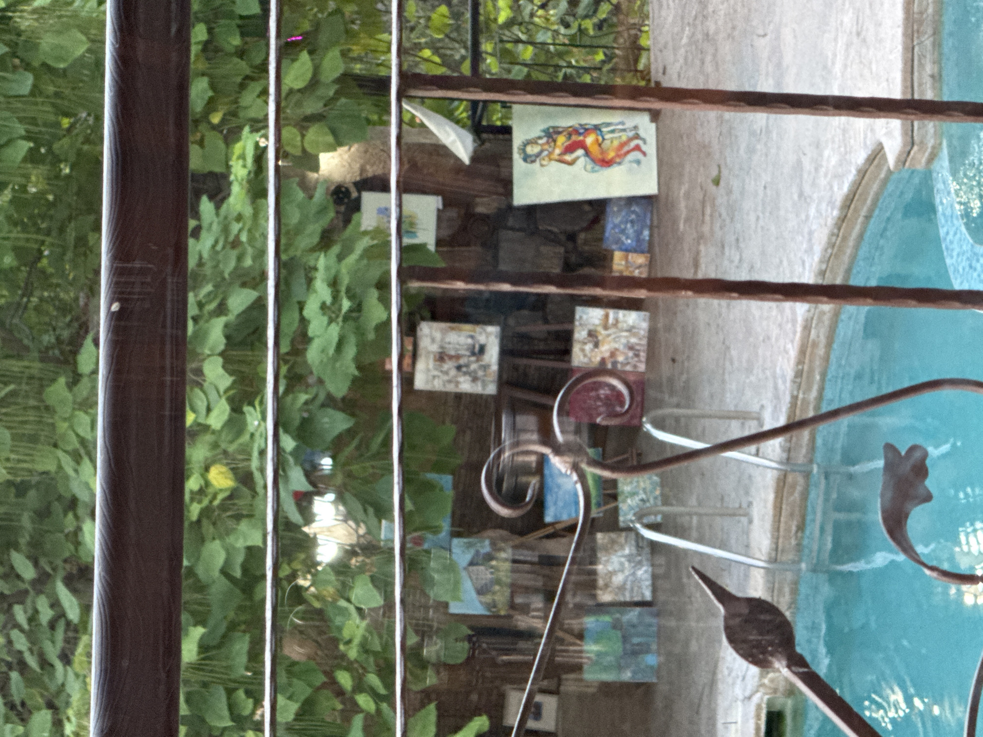 The image shows an outdoor scene with a swimming pool in the foreground and an art display in the background. The pool is filled with turquoise water and has metal railings. Behind the pool, several paintings are displayed on easels and leaning against a wall. The paintings vary in style and subject matter, with some depicting landscapes and others featuring abstract or figurative elements. Lush green foliage forms a dense backdrop, suggesting a garden or natural setting. The overall atmosphere appears to be relaxed and artistic. This scene is located in the city of Sredets, Bulgaria.