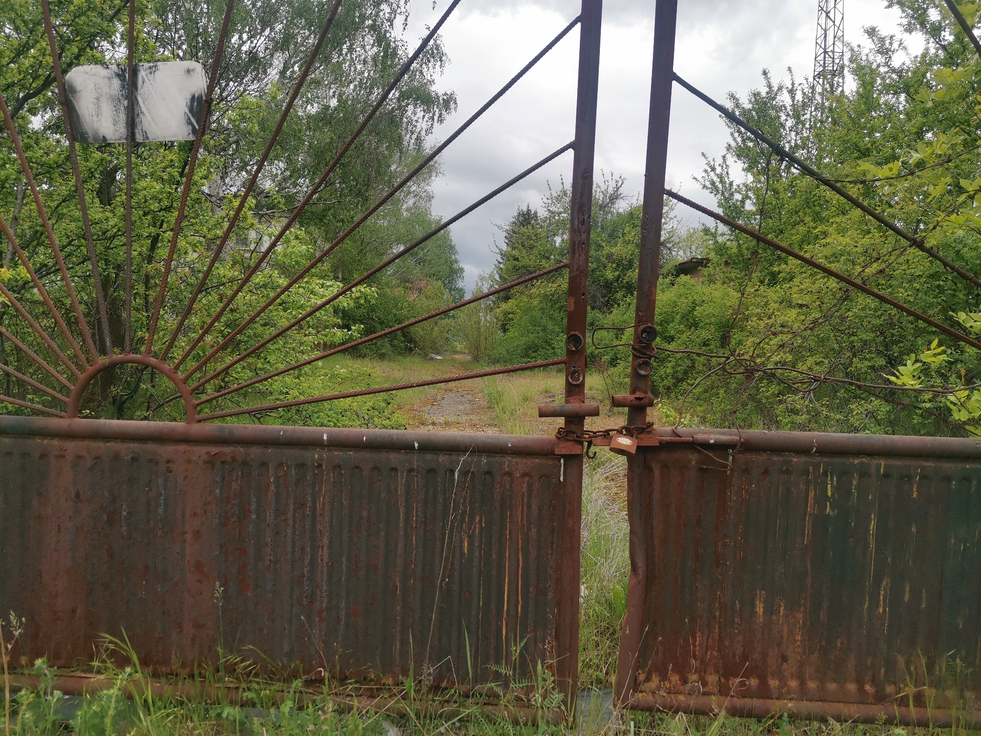 A rusty, deteriorated metal gate, slightly ajar, blocks a path overgrown with grass and vegetation. The gate's design features vertical ridged panels and a semi-circular sunburst pattern on one side. A chain and padlock secure the gate. The surrounding environment is heavily vegetated with trees and shrubs. An electrical transmission tower can be seen in the distance on the right. The location is suggested as Verinsko, Bulgaria.