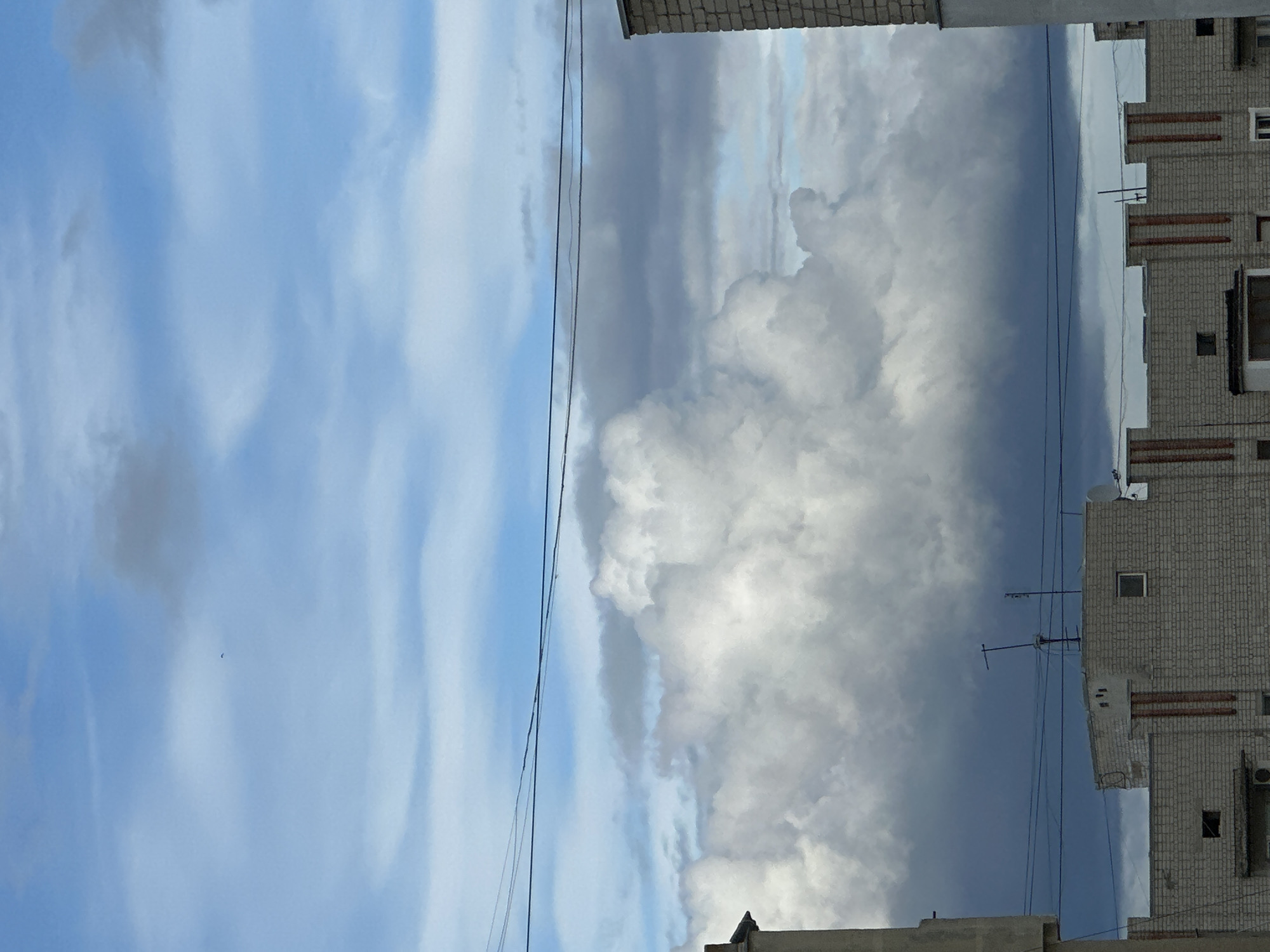 The image shows a partly cloudy sky next to a building. Electrical lines run vertically from top to bottom through the center of the image, near the break between the sky and the building. The left side of the image shows a blue sky with white streaky clouds. The right side shows a large cloud formation, appearing grey and white, beside a brick building with several windows and antennas on its roof. The location appears to be an urban area of an unknown city.