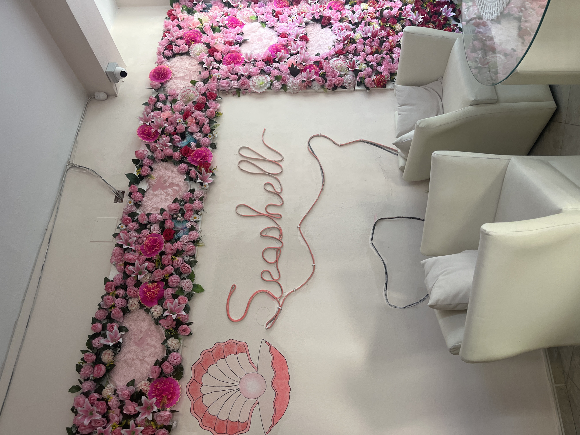 The image depicts an interior scene, likely a cafe or lounge, in Bashkia Durrës, Albania. The walls are adorned with an abundant floral arrangement in shades of pink and fuchsia, featuring various types of artificial flowers such as roses and lilies. A large drawing of a pink seashell with a pearl is visible on the left wall. To the right of the seashell drawing, the word "Seashell" is rendered in neon pink lighting, with visible wiring attached to the wall. Two plush white armchairs with matching white cushions are positioned in the foreground, facing a round glass-top table with a white knitted overlay. A security camera is mounted on the upper right wall, near the ceiling. The floor appears to be tiled. The overall aesthetic is bright, feminine, and decorative.
