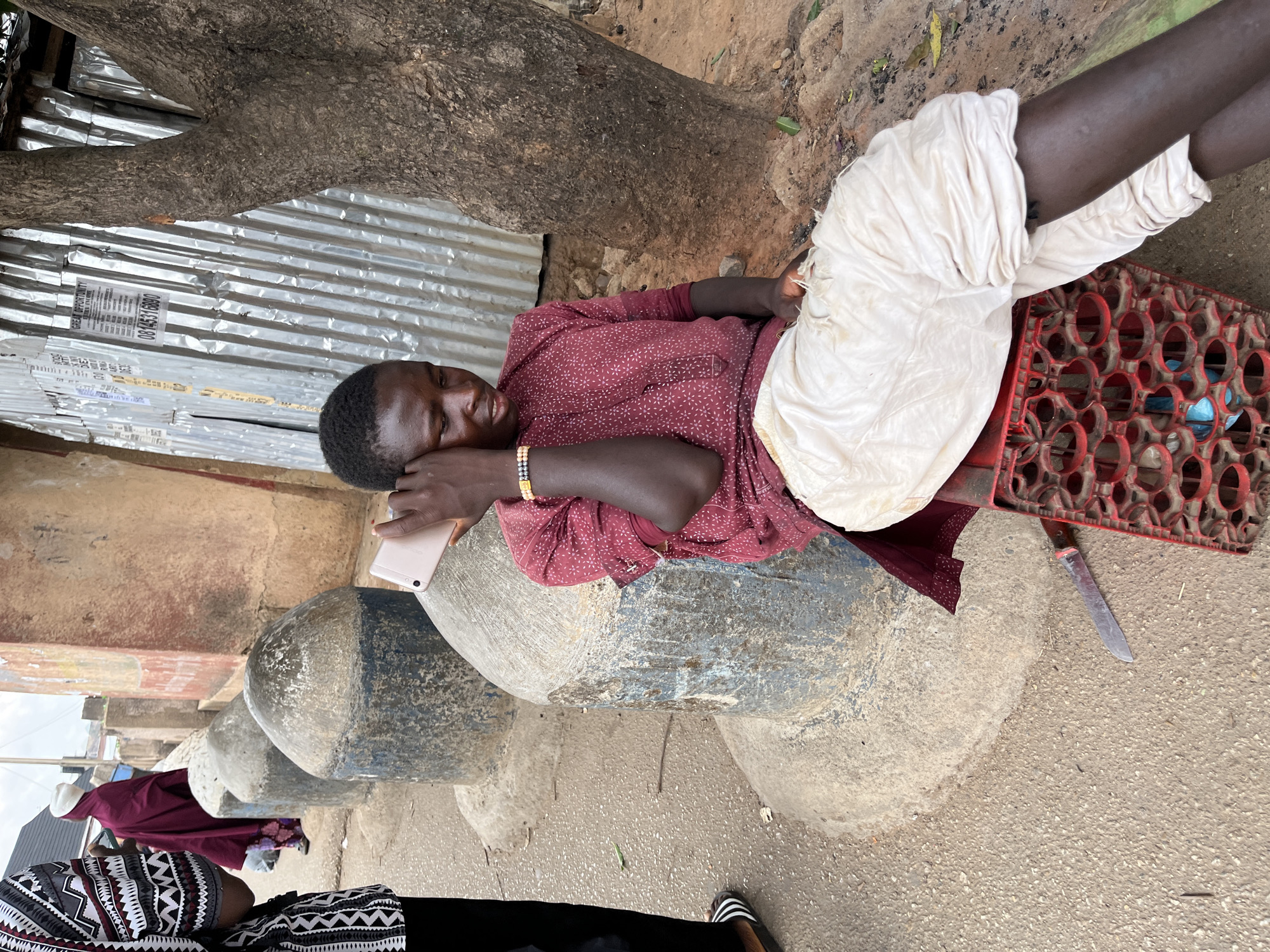 A man is reclining on a row of concrete structures in what appears to be a public space. He wears a maroon shirt with a small pattern, light-colored trousers, and a beaded bracelet. He is holding a mobile phone to his ear. Beside him, a red plastic crate sits on the ground, along with a knife. A corrugated metal wall and a tree trunk are visible in the background. The scene is believed to be in the city of Jos, Nigeria.