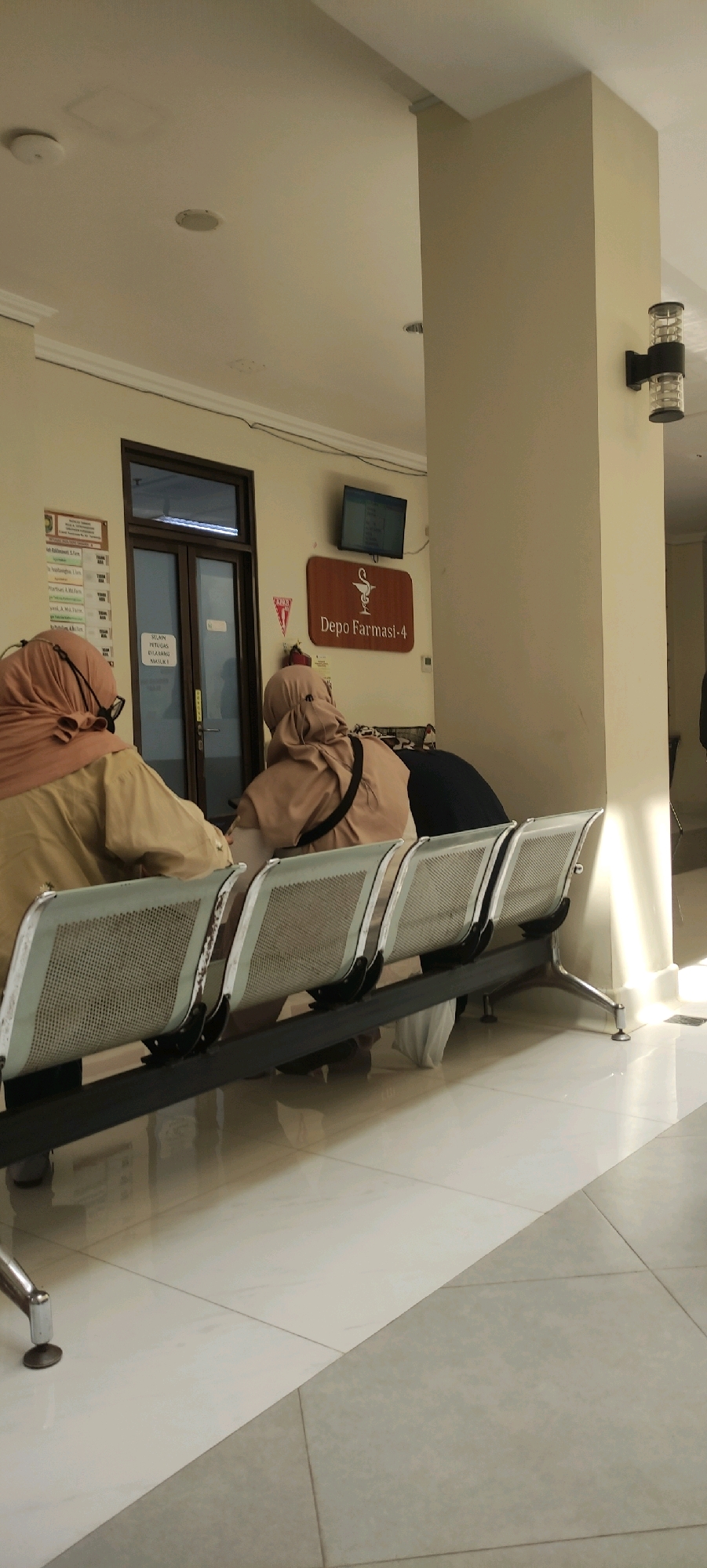 The image depicts a waiting area, likely a clinic or pharmacy, with three individuals seated on a bench of four chairs. The individuals are wearing hijabs and appear to be waiting. The environment includes a tiled floor, a light-colored wall, a doorway with glass doors, a sign reading "Depo Farmasi-4" with a caduceus symbol, a small television screen mounted on the wall, and an outdoor wall lamp on a pillar. The scene is well-lit with natural light coming from the right. The location is identified as Purworejo, Indonesia.