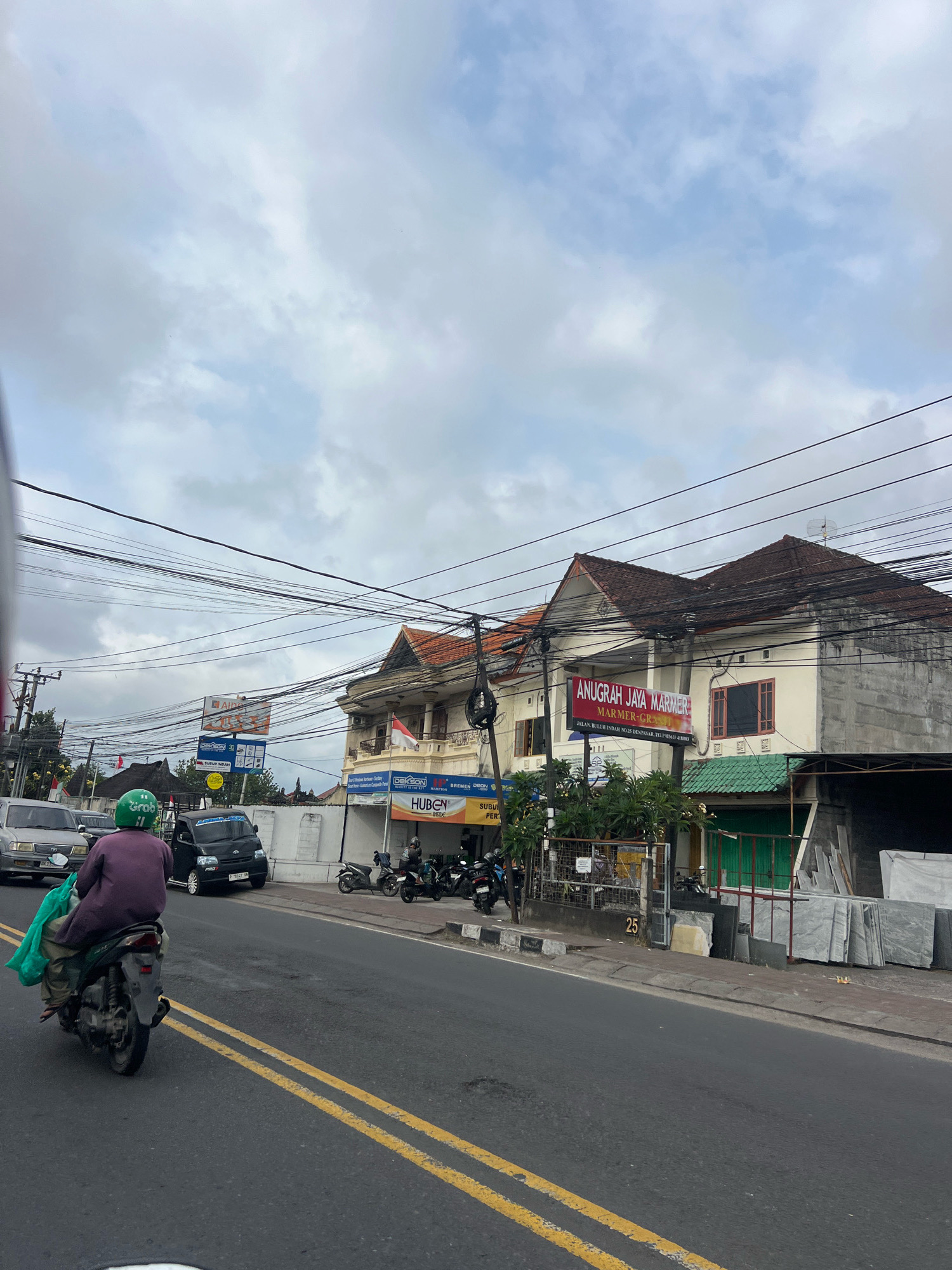 The image depicts a street scene in Denpasar, Indonesia, with a focus on a section of road lined with buildings. In the foreground, a person on a scooter, wearing a green helmet with a "Grab" logo, is riding away from the viewer. They are carrying a green bag over their shoulder. The scooter is on the right side of the road, which has double yellow lines marking the lanes.
On the right side of the road, there are several buildings. One building displays a sign that reads "ANUGRAH JAYA MARMER" and "MARMER-GRANIT". Another sign below it provides an address: "JLN. BULUH INDAH NO.25 DENPASAR". Adjacent to this building is a shop with a green awning and stacked slabs of marble or granite outside. Further down the street, there are more parked motorcycles.
The sky is overcast with a mix of clouds and blue sky. Overhead, numerous electrical wires crisscross the scene, creating a complex network against the sky. Various vehicles, including cars and other scooters, are visible further down the road, suggesting it is a moderately busy urban area. The overall impression is that of a typical street in Denpasar during the daytime.