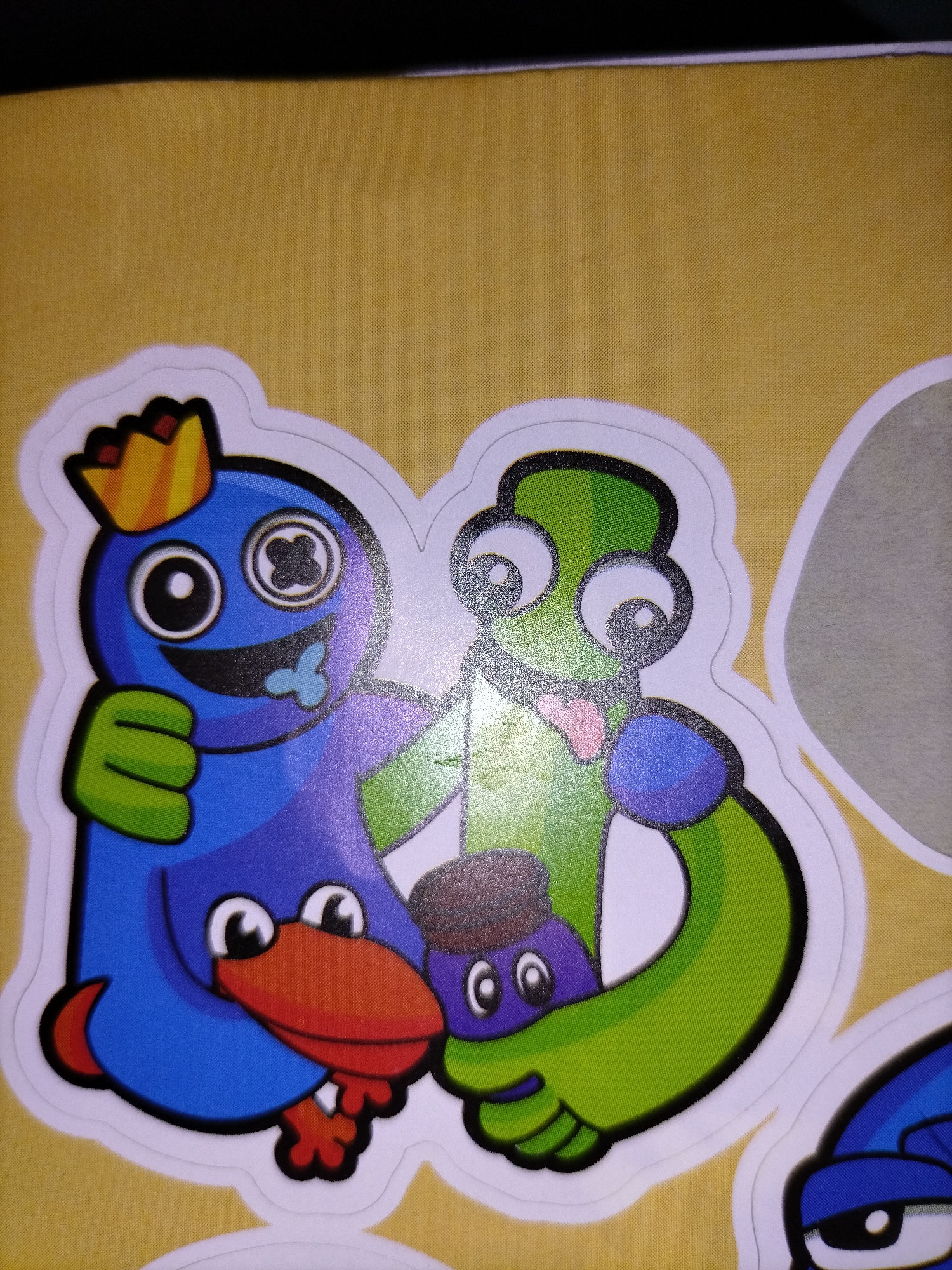 This media file features two cartoon characters on a yellow background, possibly a sticker or a piece of artwork. The character on the left is blue, wearing a golden crown, and has a large red beak. It is embracing the character on the right, which is green and has a more elongated body. Both characters have large, cartoonish eyes. A smaller, purple character with two eyes is visible between them, seemingly being hugged by both. The overall mood appears to be one of friendship or affection, as suggested by the embraces. The image is cropped, and part of another blue character with prominent eyes is visible on the right side. There is no visible text. The setting is a generic yellow surface, possibly a table or wall, with no discernible location cues beyond the provided context of Smalyavichy, Belarus.