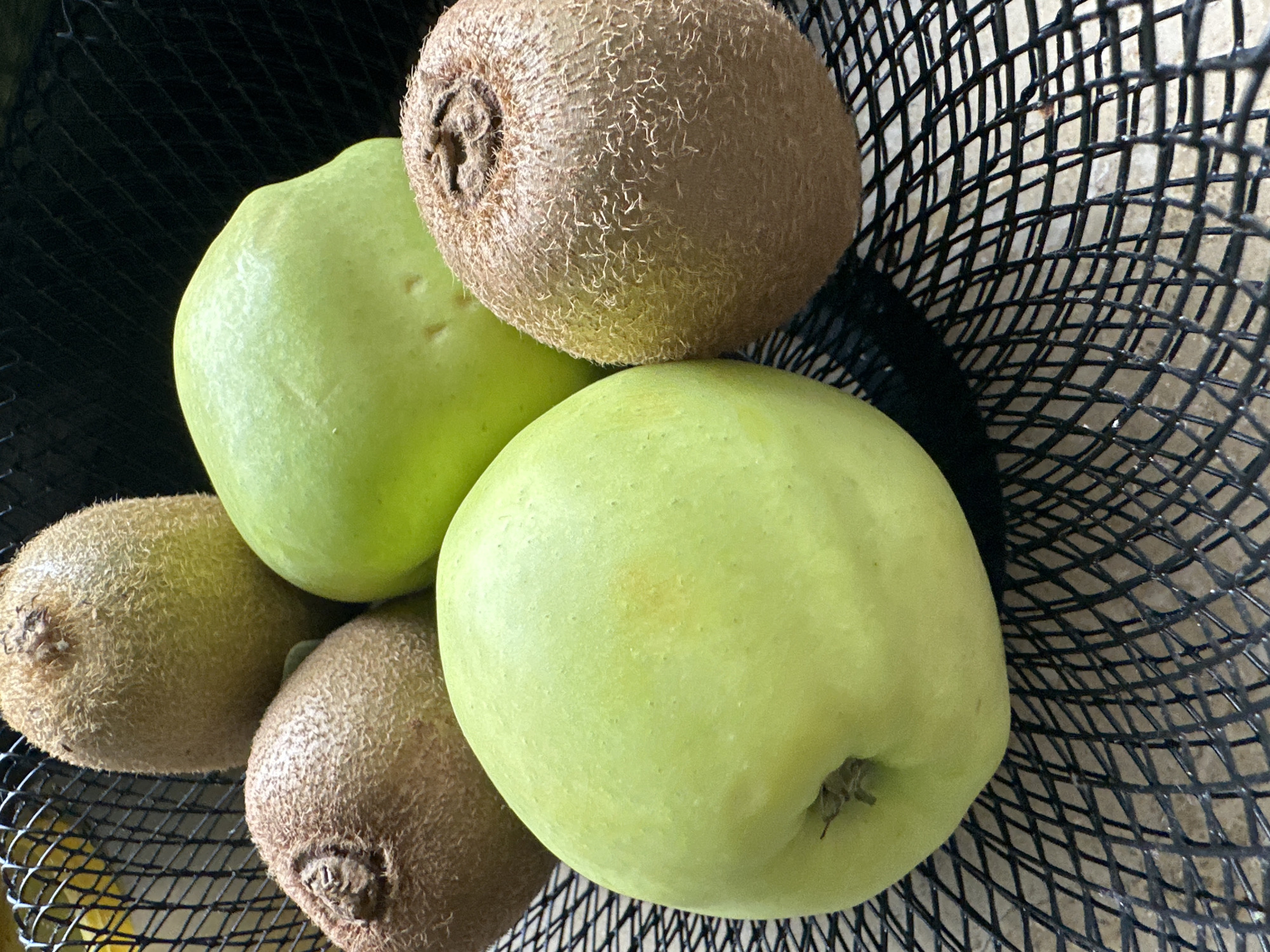 This is a close-up, overhead shot of a black wire basket filled with several pieces of fruit. The subjects are two green apples and three kiwis. The apples are smooth and round, with a light green color. The kiwis are oval-shaped and covered in a fuzzy brown skin. The basket's mesh pattern is clearly visible and forms a grid-like background behind the fruit. The lighting suggests it's daytime, casting soft shadows. There are no people or discernible text in the image. The overall scene is a simple still life of fruit in a container, likely in a kitchen or on a counter. There are no cues to indicate the specific location within Krefeld, Germany, other than it is a general setting.