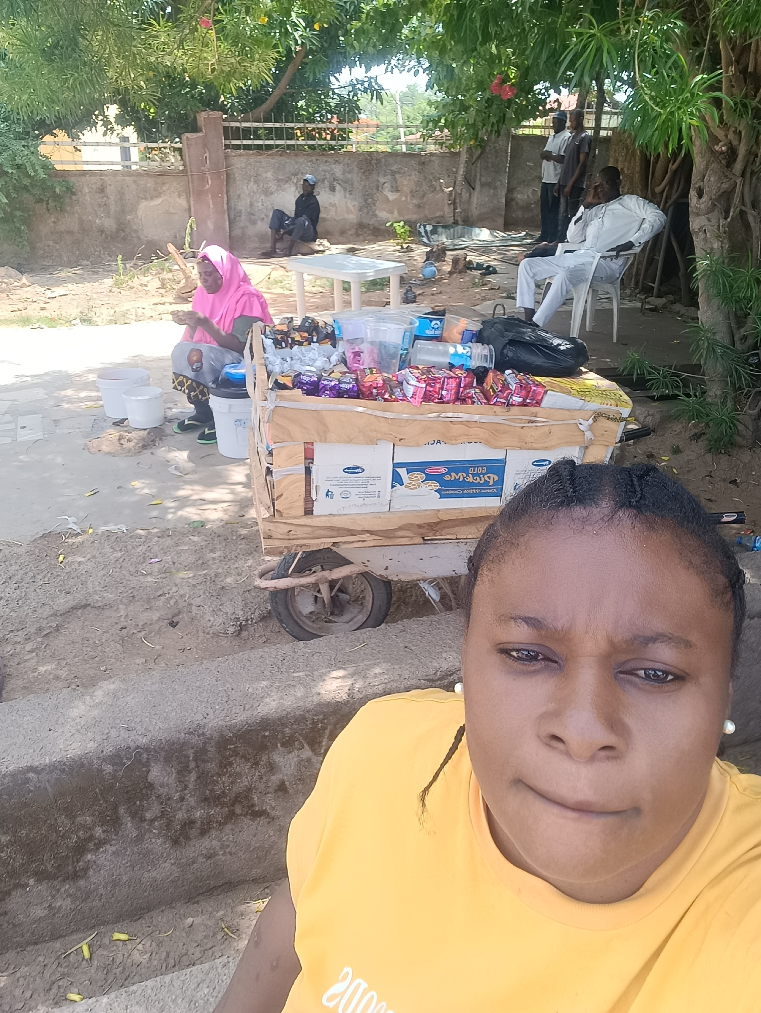 The photograph appears to be a selfie taken near a street vending setup, potentially located in Jos, Nigeria. A woman wearing a yellow t-shirt is in the foreground. Behind her, there is a wooden cart laden with various packaged snacks, cookies, and a black plastic bag. To the left of the cart, a woman in a pink hijab sits on a small stool. Several white buckets are visible near her. Further back, several men are visible; one is sitting on a white plastic chair. The scene is partially shaded by trees. A fence and some buildings are visible in the background.