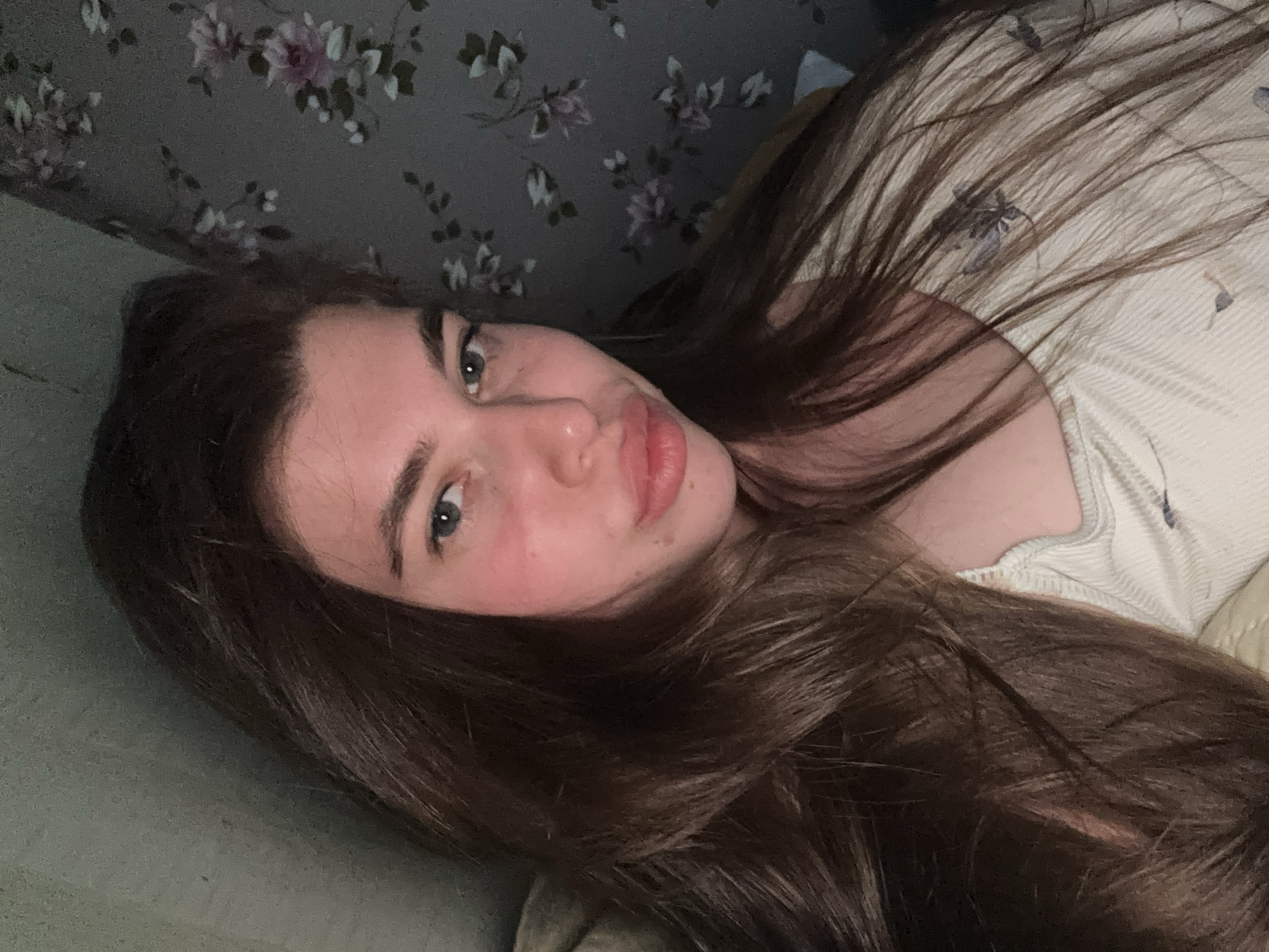 A young woman with long, light brown hair lies on a surface, possibly a bed or sofa, facing the camera. She has fair skin, light eyes, and slightly pursed lips. Behind her is a wall with a floral pattern in muted colors. The lighting is soft and somewhat dim. The location cannot be determined as Мещёры, Russia based on the available information.
