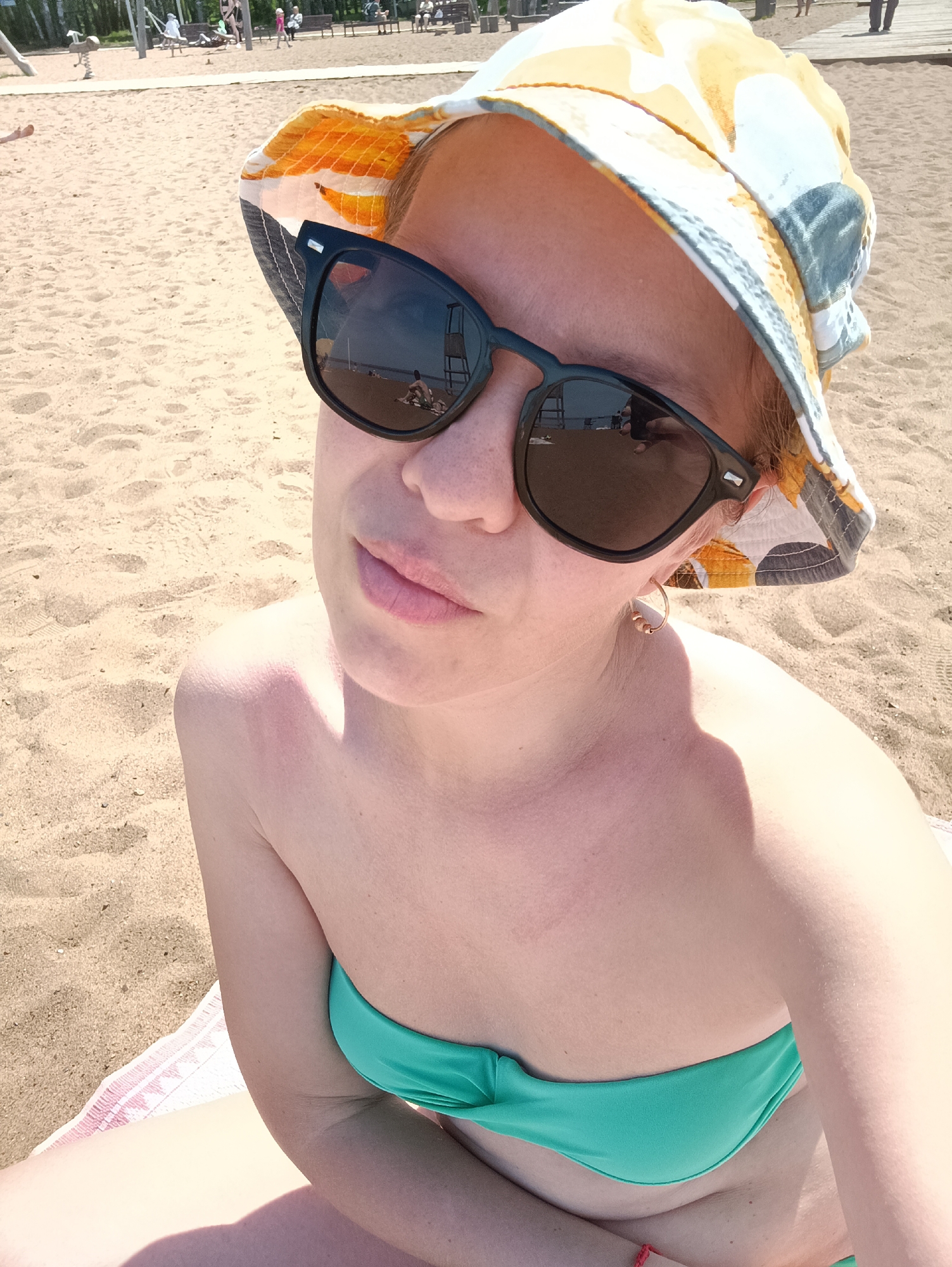 A medium-angle eye-level shot depicts a woman at a beach in Chaikovsky, Russia. The woman is wearing a green bikini top and a hat with a pattern of yellow, orange, and grey-blue shades. She is also wearing sunglasses that reflect the scene of the beach, including the lifeguard tower. The woman has light skin. The beach consists of light brown sand with some footprints. In the background, there are people, beach equipment, and trees, indicating a public area. The lighting appears to be sunny, with shadows and bright highlights on the sand and the woman's skin. A portion of a towel is visible in the lower-left corner.
