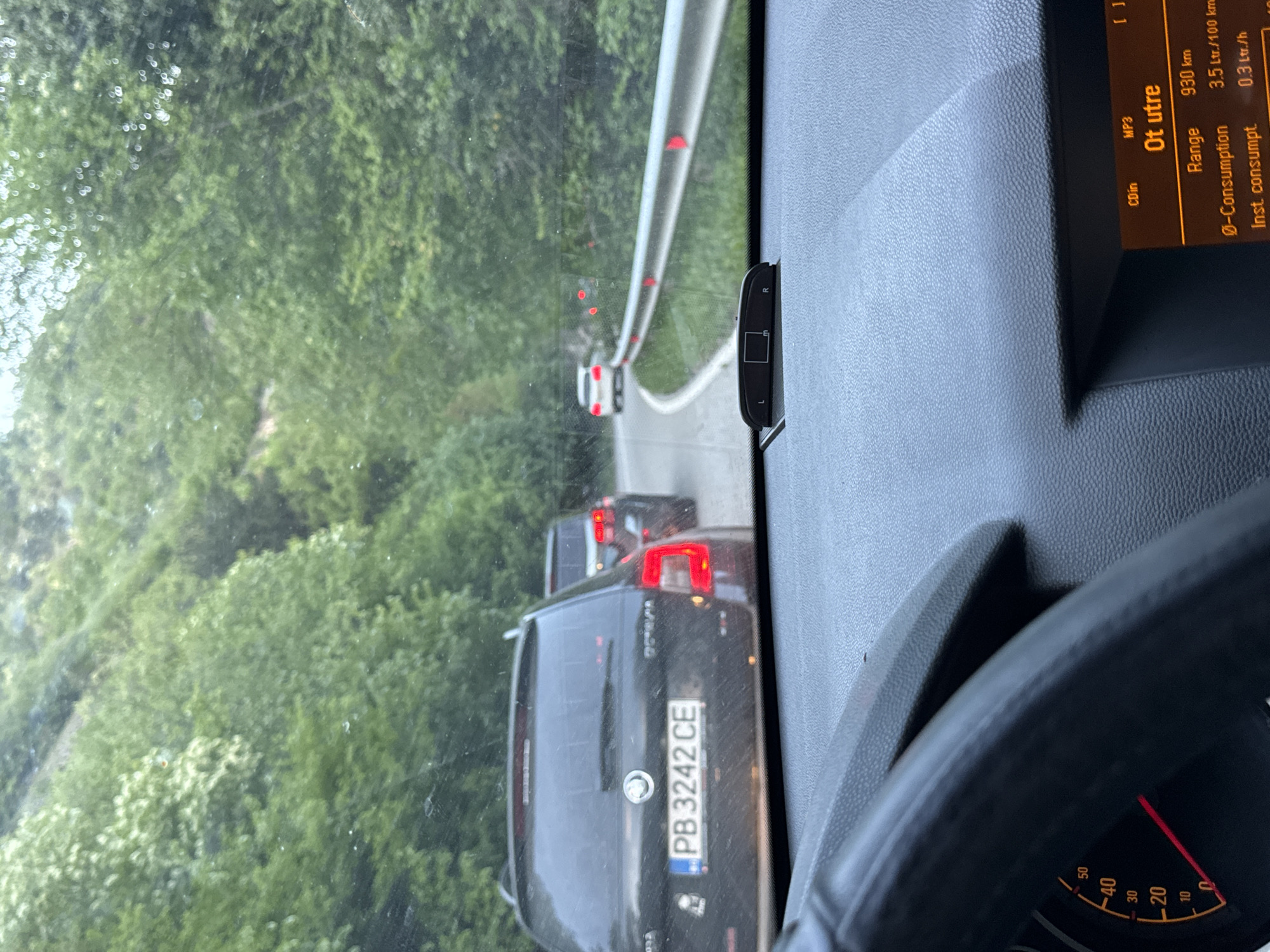 The image, taken from inside a vehicle in what appears to be heavy traffic near Бачковски манастир, Bulgaria, shows the dashboard, steering wheel, and the road ahead through the windshield. The dashboard display reads "Ot utre," with a range of 930 km and a consumption rate of 3.5 Ltr/100 km. A black car with the license plate "PB 3242 CE" is visible in front, followed by at least two more vehicles. A guardrail borders the road, and a dense forest lines the roadside.