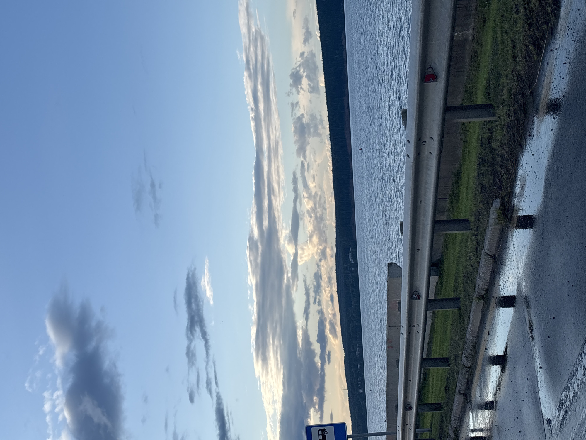 A roadside scene along a body of water features a guardrail and a wet asphalt surface. A blue sign with a white bus icon is visible on the left. The water is calm and stretches to a tree-lined horizon. The sky is partly cloudy with a mix of blue and grey clouds, suggesting it might be late afternoon or early morning. The scene appears to be located in the city of Chaikovsky, Russia, based on visual cues such as the landscape and typical roadside infrastructure.