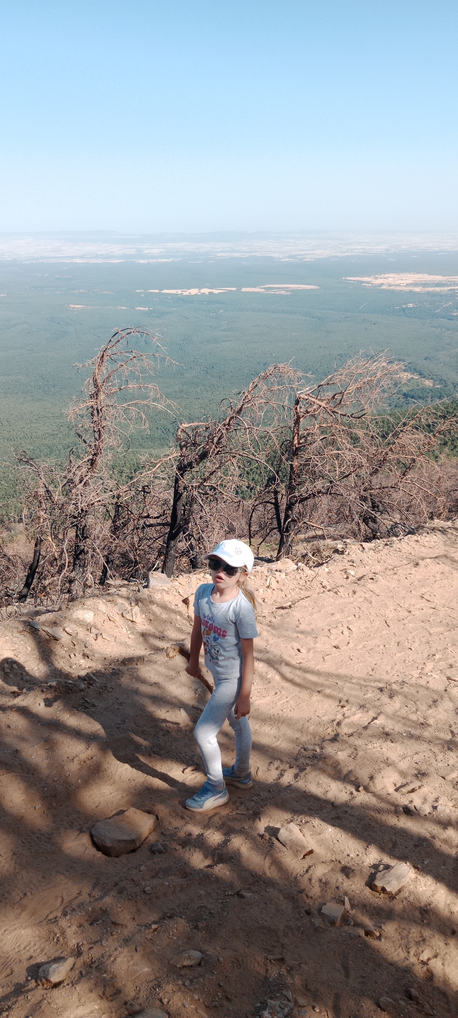 The image shows a young girl standing on a dirt path on a hillside. She is wearing a white cap, sunglasses, a grey t-shirt, and grey leggings. The girl is holding a stick in her right hand. Behind her, several dead, burnt trees are visible. The ground is covered in sand, small rocks, and dry vegetation. In the background, a vast expanse of green forest stretches out to the horizon, meeting a hazy blue sky. The scene suggests a location in a natural, possibly mountainous area, with evidence of past fire damage to the trees. The girl appears to be exploring or hiking. The city of Cerezo de Arriba, Spain is known for its mountainous terrain and pine forests, which can be susceptible to wildfires.