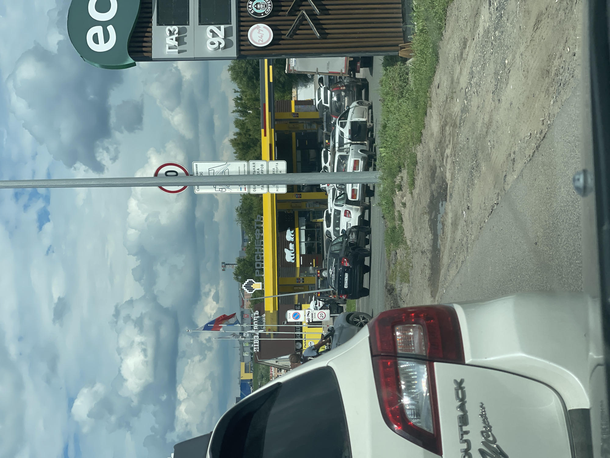 A line of cars is queued at a gas station in Samara, Russia, under a partly cloudy sky. A white Subaru Outback is in the foreground, its rear visible. Beyond it, a yellow gas station with the logo "ПЯТЬ ДОРОГ" and "РОСНЕФТЬ" is manned by an attendant in a gray shirt. Cars are filling up at the pumps, and a sign indicates "ГАЗ" and "92" fuel prices. Another sign shows "ec" in large green letters. To the right, a "STARBUCKS" logo is visible. Russian flags fly in the background. The road surface is partially paved and partially dirt, with patches of grass.