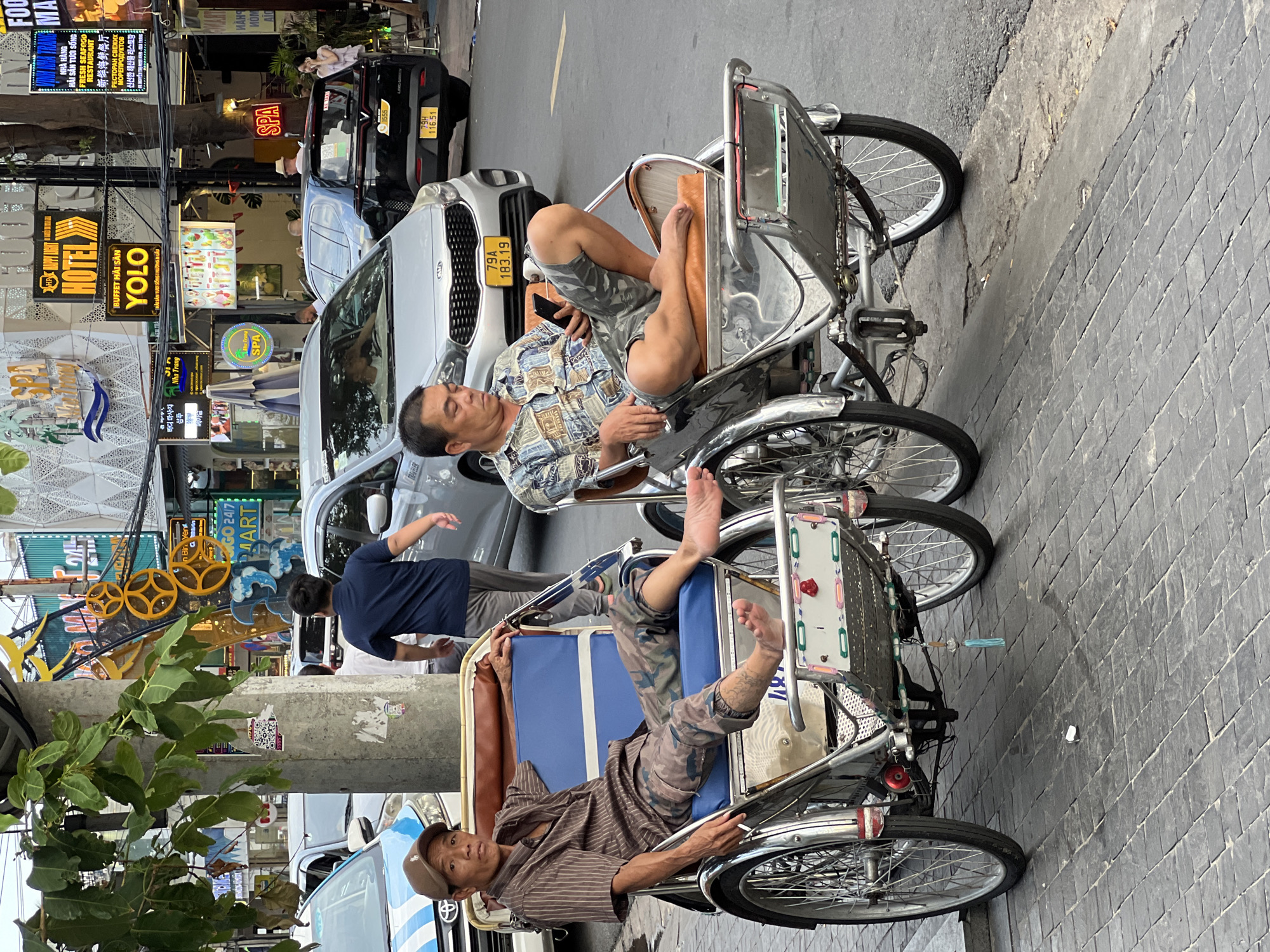 Two men sit in cyclos, a common form of transportation in Nha Trang, Vietnam, waiting for customers on a paved sidewalk adjacent to a busy street. The man on the left, wearing a brown striped shirt and camouflage pants, sits with his legs extended, looking directly at the camera. He has tattoos on his right leg. The man on the right, in a patterned shirt and camouflage shorts, is looking at his phone and has his legs crossed. Both men are barefoot. The cyclos have large spoked wheels and blue and brown seating. In the background, cars and other vehicles are visible on the street, along with various businesses with illuminated signs, including "HOTEL," "SPA," and "YOLO." The buildings are modern, with some displaying Korean text. The scene suggests a typical day in a tourist area of the city.