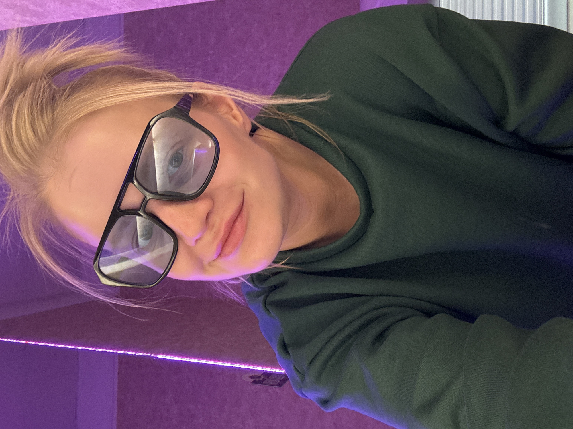 A blond woman with glasses poses in front of a purple wall illuminated by a neon light. She wears a dark green sweatshirt. The location cannot be determined as Saint Petersburg, Russia based on the information in the picture.