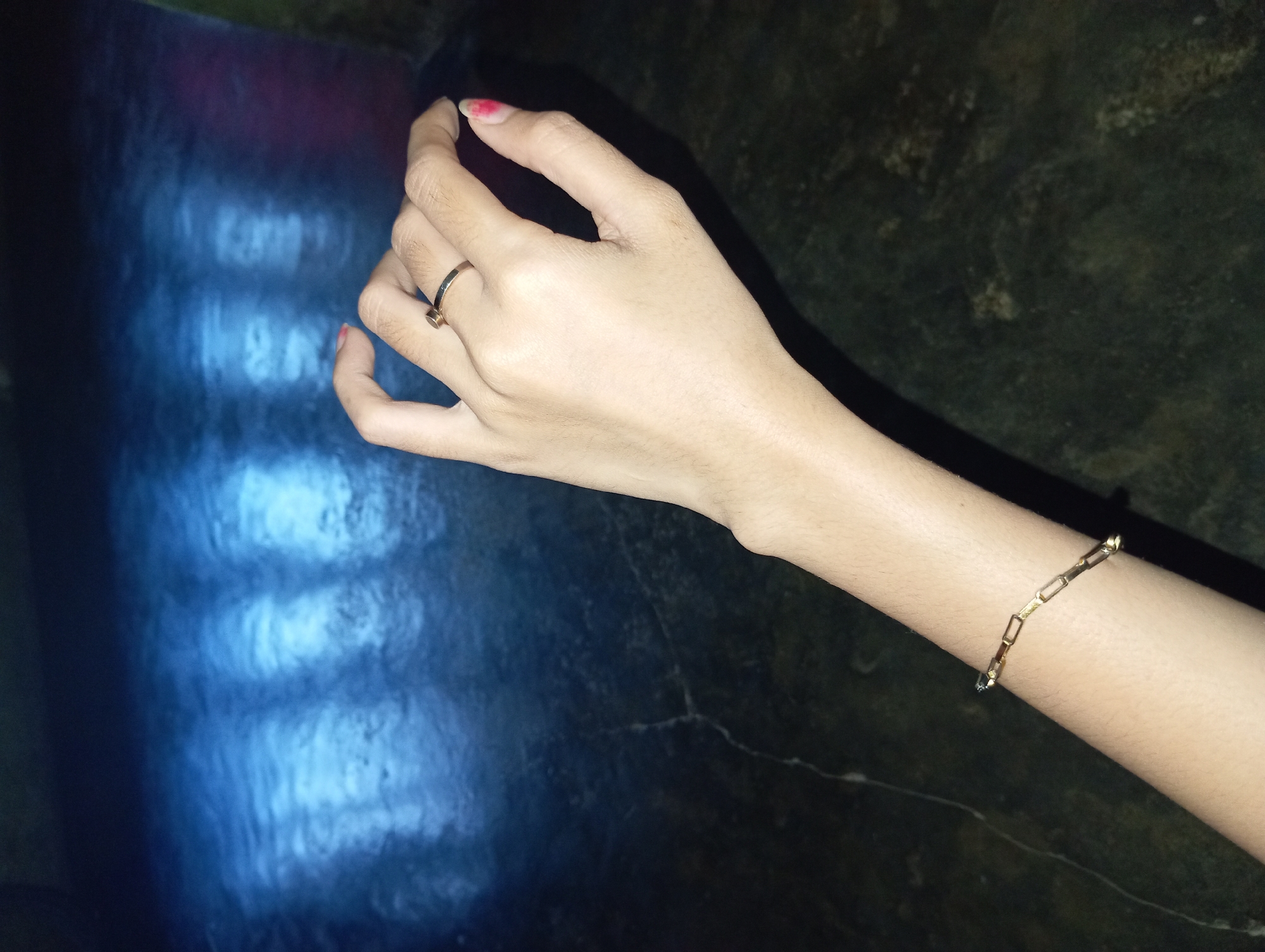 An overhead, close-up shot captures a left arm adorned with a gold chain bracelet and a gold ring on the middle finger. The subject's arm and hand are positioned against a dark, textured surface, possibly a countertop or floor, with subtle cracks visible. The background is dominated by a dark blue, abstract blur of light, reflecting what appear to be three vertical light sources. The lighting suggests an indoor setting, potentially at night. The scene is static, with no discernible action or interaction occurring. The location is identified as Rejosari, Indonesia, although no specific landmarks or context within the city are evident from the image alone.