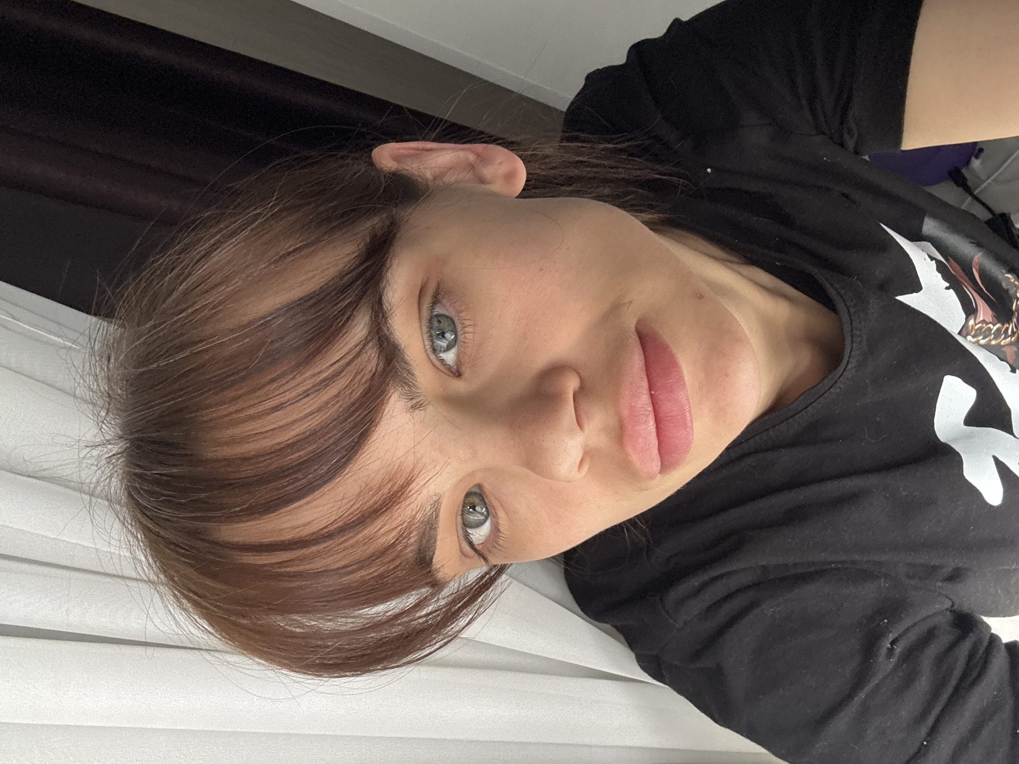 This media file presents a close-up, tilted portrait of an individual, likely female, with fair skin and piercing blue-green or hazel eyes that gaze directly towards the viewer. Her light brown hair, styled with full, straight bangs across her forehead and longer strands framing her face, suggests a natural and relaxed look. Her lips are full and slightly parted, showing a natural pink tint, and her expression is neutral and calm, conveying a sense of quiet introspection.
She is wearing a simple black t-shirt, which features a subtle white graphic design and a gold-colored chain detail visible on the chest area, hinting at a larger print. A minor blemish is discernible on her lower cheek/chin, and a faint area of redness appears on her neck.
The background elements suggest an indoor setting. Behind her head, a dark, indistinct area is visible, possibly a curtain or dark piece of furniture. To the lower left, a white, horizontally textured surface, resembling window blinds or a similar paneling, can be seen. Below her body, there is a soft, light-colored fabric surface, possibly a bed or couch, with a hint of a purple object in the far background.
The image captures a casual moment, likely a self-portrait, in what appears to be a home environment. The lighting suggests it was taken during daytime hours, with soft, natural illumination. The photo's context places it in Rostov-on-Don, Russia. No readable text is visible within the frame.