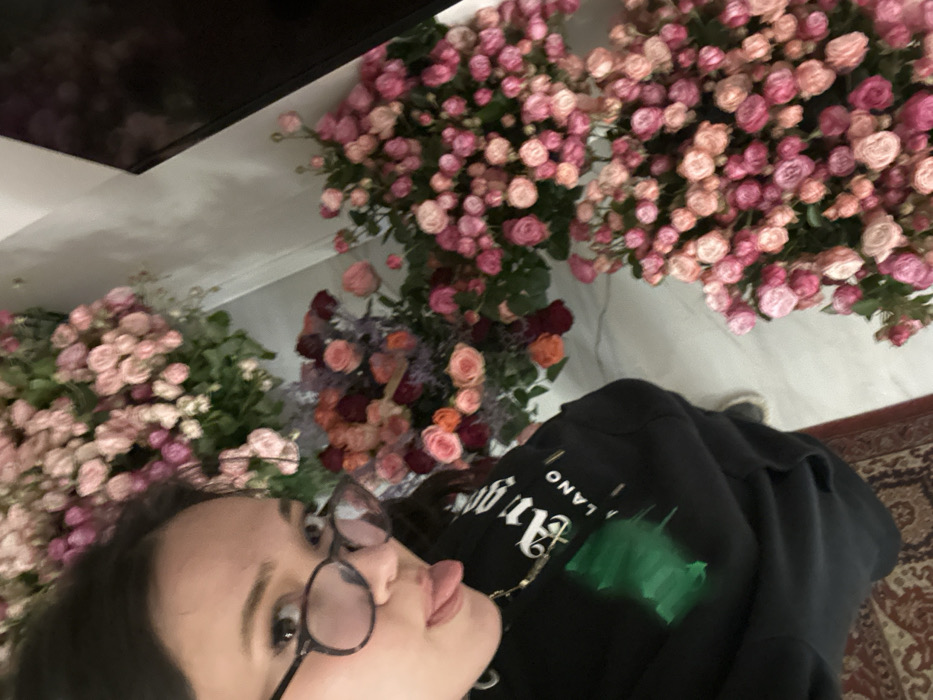 This image depicts a female individual taking a selfie in what appears to be a floral shop or display area in Yerevan, Armenia. The subject's face is visible in the upper left, characterized by dark hair, dark eyes, and tortoiseshell-frame glasses. Her tongue is partially extended from her mouth. She is wearing a black hooded sweatshirt with white text, including "MILANO," and a green graphic on the front. A thin, gold-toned chain necklace is also visible. The background is densely filled with extensive floral arrangements, primarily composed of miniature roses in varying shades of pink, light pink, and reddish-orange. The floor consists of a light-colored, reflective surface, possibly marble or tile, with a segment of a patterned red and brown rug visible in the lower left. A dark, reflective rectangular object is partially visible on the upper right edge of the frame.