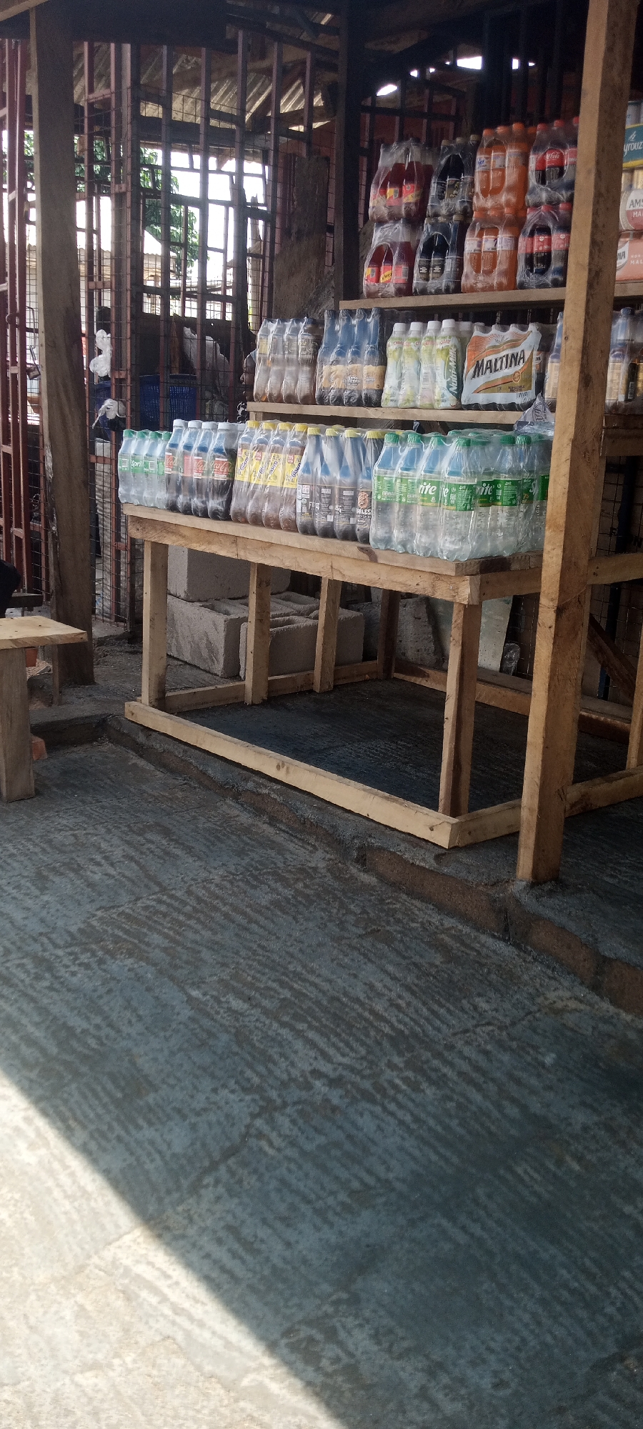 In Jalingo, Nigeria, a roadside stall displays a variety of bottled beverages on wooden shelves. The stall is constructed from a mix of wood and metal, with a corrugated metal roof. In the foreground, the ground is covered with a dark, textured material, possibly concrete or asphalt, with some areas appearing wet or recently poured. A wooden bench or table is visible on the left.
The shelves are stocked with numerous bottles of soft drinks, many of which are wrapped in plastic. On the upper shelves, stacks of plastic bottles containing orange-colored and dark beverages are visible, along with some bottles of what appears to be Maltina. The lower shelf holds rows of clear plastic bottles containing clear or light-colored liquids, including Sprite. A few stacks of cement blocks are placed beneath the lower shelf.
In the background, a person can be seen standing near the rear of the stall, partially obscured by the structure. Beyond the stall, a building with metal bars and a roof is visible, suggesting an urban or semi-urban environment. The overall scene depicts a typical small business operation in Nigeria, likely a beverage vendor.