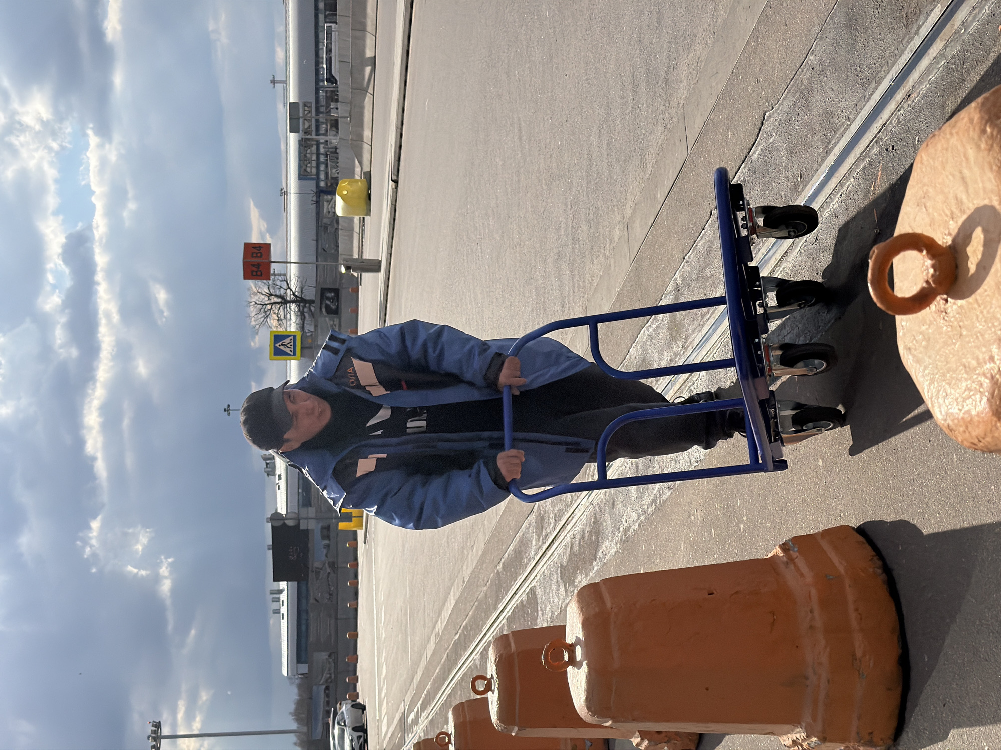 An outdoor, eye-level shot depicts a man standing on a paved surface at what appears to be an airport in Domodedovo, Russia. The man, positioned in the foreground, is wearing a blue work jacket with "ОДА" (ODA) partially visible on the shoulder, a black hoodie, and a dark cap. He holds onto a blue metal hand truck or dolly with four small wheels. His expression is neutral, with a slight hint of a smile.
To his right, a series of orange concrete barriers, each topped with a metal ring, line the edge of the paved area, extending into the distance. The ground is a light gray asphalt or concrete. The sky above is partly cloudy with significant patches of blue.
In the mid-ground and background, typical airport infrastructure is visible, including distant buildings (likely terminals or hangars), light poles, and a yellow marker or cone. Two notable signs are present: an orange rectangular sign displaying "B4 B4" in white text, and beneath it, a yellow triangular warning sign with a black pedestrian symbol, indicating a pedestrian crossing. A bare tree stands in the mid-ground, and a white car is faintly seen further in the distance. The scene is brightly lit by natural daylight, casting distinct shadows and suggesting a sunny or partly sunny day.