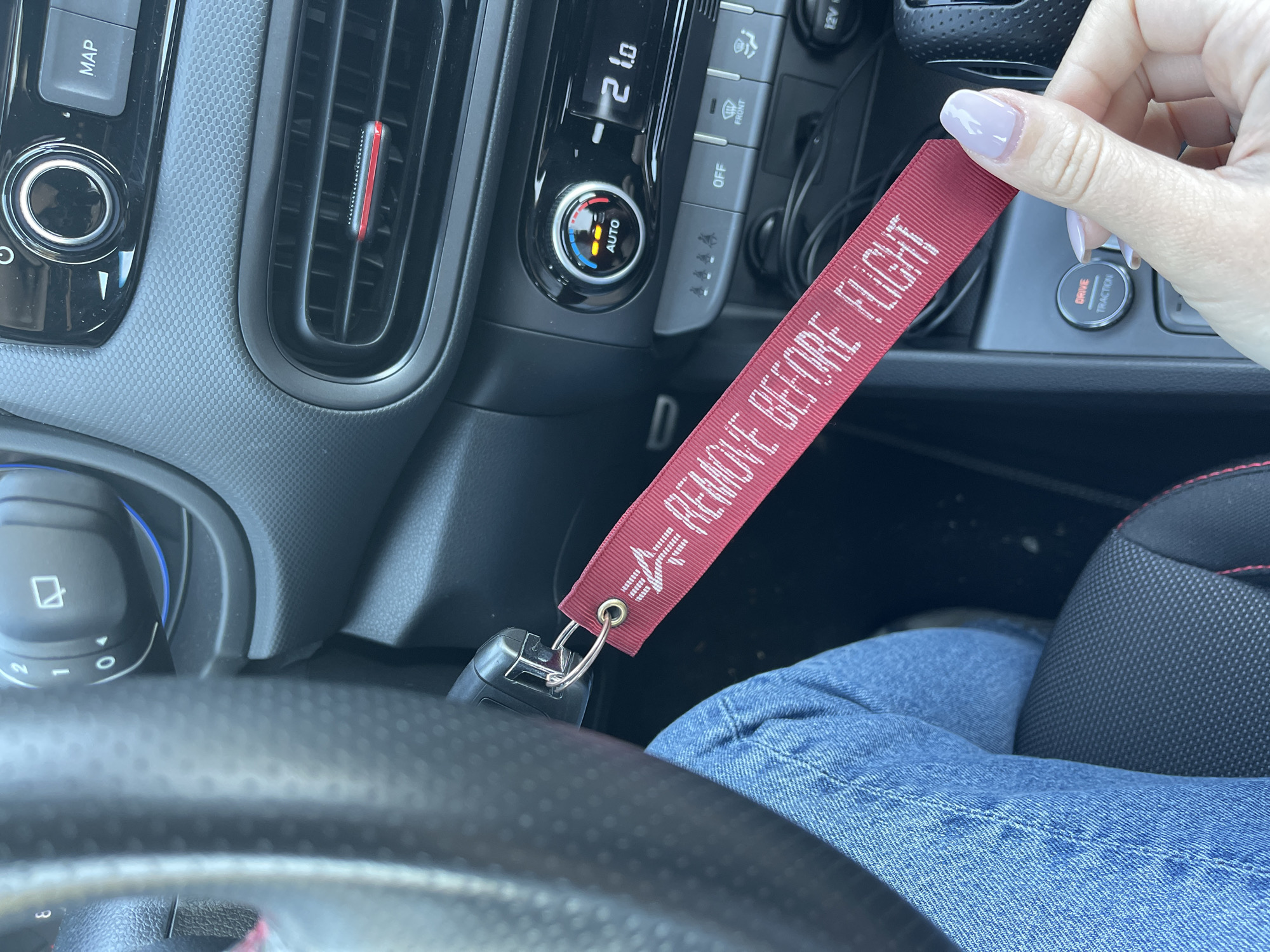 The image shows the interior of a car, with a focus on the dashboard and steering wheel. A hand holds a red keychain with the text "REMOVE BEFORE FLIGHT". The dashboard features climate controls, including a digital display showing "21.0" and an "AUTO" button. There are also buttons for defrosting and other functions. A map button is visible on the left side of the dashboard. The steering wheel is black. The person is wearing blue jeans. The location is unknown, and there is no indication of Sofia, Bulgaria.