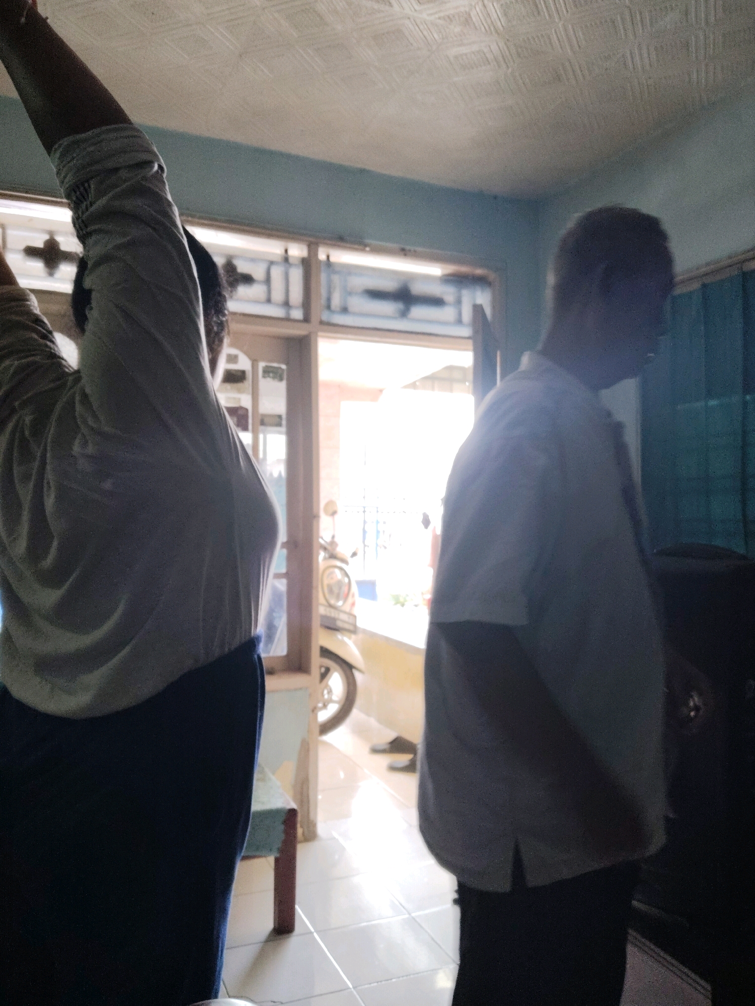 In Subang, Indonesia, a dimly lit indoor scene features two individuals. On the left, a person is seen from the side, with their right arm raised and wearing a grey long-sleeved shirt and dark trousers. Their face is obscured. To the right, an older male is captured in profile, facing away from the camera. He is wearing a light-colored short-sleeved shirt and dark trousers. Behind them, a doorway leads to an outdoor area where a scooter is partially visible. The room has a textured ceiling and pale blue walls. A dark object, possibly a piece of furniture, stands to the right of the male. The flooring appears to be tiled.
