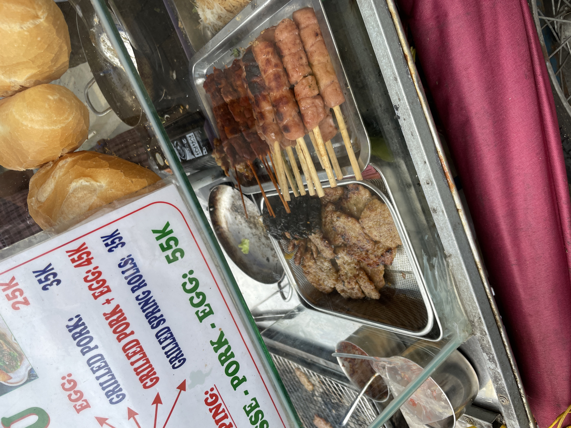 In a street-side food stall in Thủ Đức, Vietnam, a menu displays prices for grilled food items. The menu lists "EGG: 25K", "GRILLED PORK: 35K", "GRILLED PORK + EGG: 45K", "GRILLED SPRING ROLLS: 35K", and "SSE - PORK - EGG: 55K". In the foreground, a metal tray holds several skewers of grilled meat, some of which appear to be pork. Another tray contains grilled pork slices and a dark, possibly fermented, food item. In the background, three large bread rolls are visible, suggestive of banh mi. The stall appears to be open-air, with a glass display case protecting the food. The overall scene depicts a typical street food vendor preparing and selling grilled dishes.