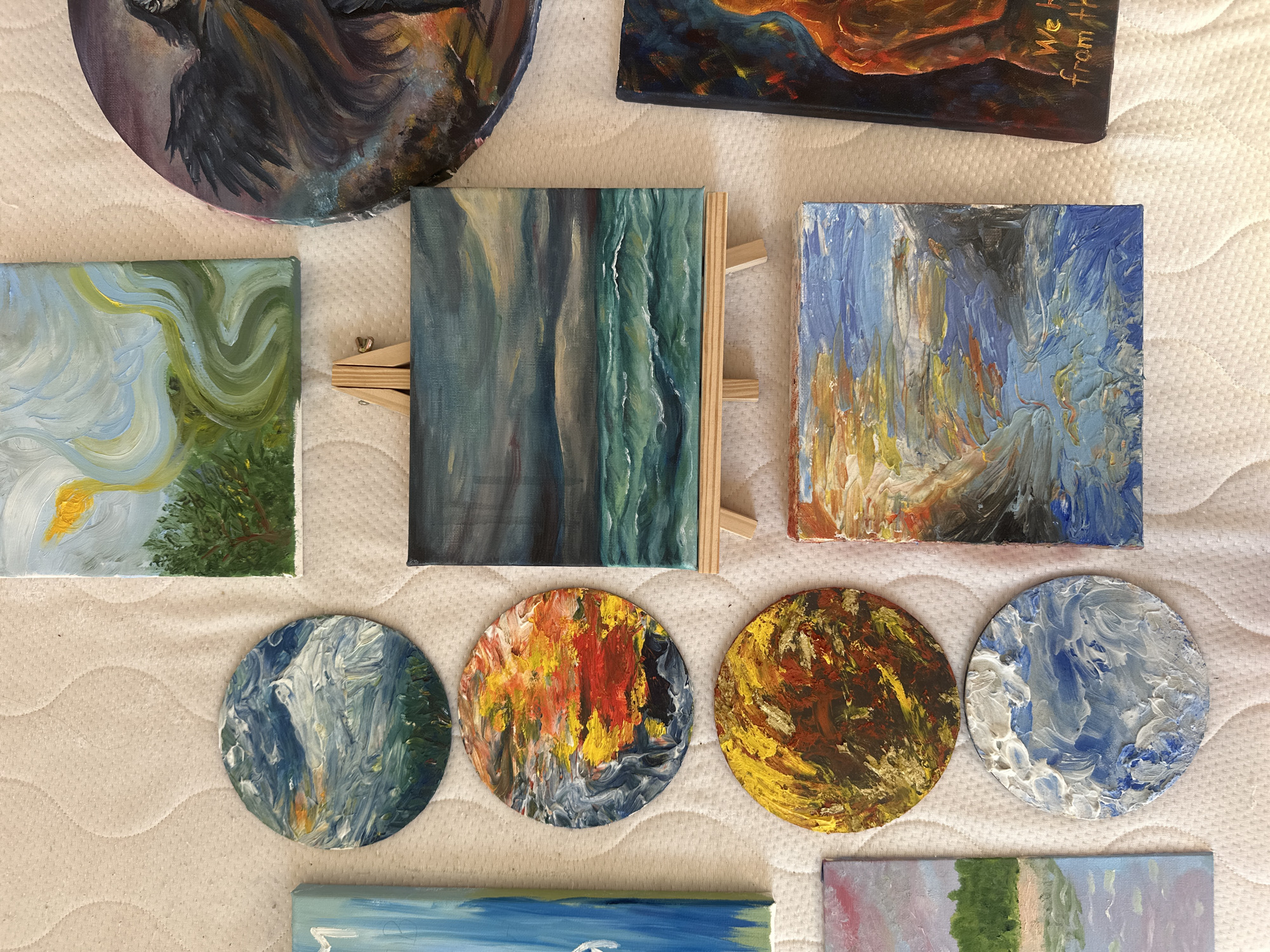 The image displays a collection of small, colorful paintings arranged on a white, textured surface, possibly a bedspread or a canvas. The paintings are varied in size and shape, including circular and rectangular canvases, with some placed on a miniature easel. The subjects appear to be abstract landscapes or seascapes, rendered in thick, impasto brushstrokes. Some of the circular paintings feature vibrant oranges, yellows, and reds, suggestive of sunsets or fiery elements, while others are dominated by blues and whites, evoking water or clouds. A rectangular painting on an easel depicts a stormy seascape with dark, ominous clouds over choppy, teal-colored water. Another circular painting shows a figure with dark, feathered wings, possibly an angel or a mythological creature, in dark, moody tones. To the right of the easel painting is a rectangular artwork featuring a person's torso in warm, fiery colors, with handwritten text at the bottom that reads "We h from th". A thin, vertical blue painting is partially visible on the left side of the image, and below it is a landscape with green trees and a body of water. Another rectangular painting on the bottom left shows a serene landscape with a body of water and distant trees. The overall impression is of an artist's collection of works in progress or a display of finished pieces. The scene does not contain any identifiable landmarks or elements that would suggest a specific location in Prague, Czechia.