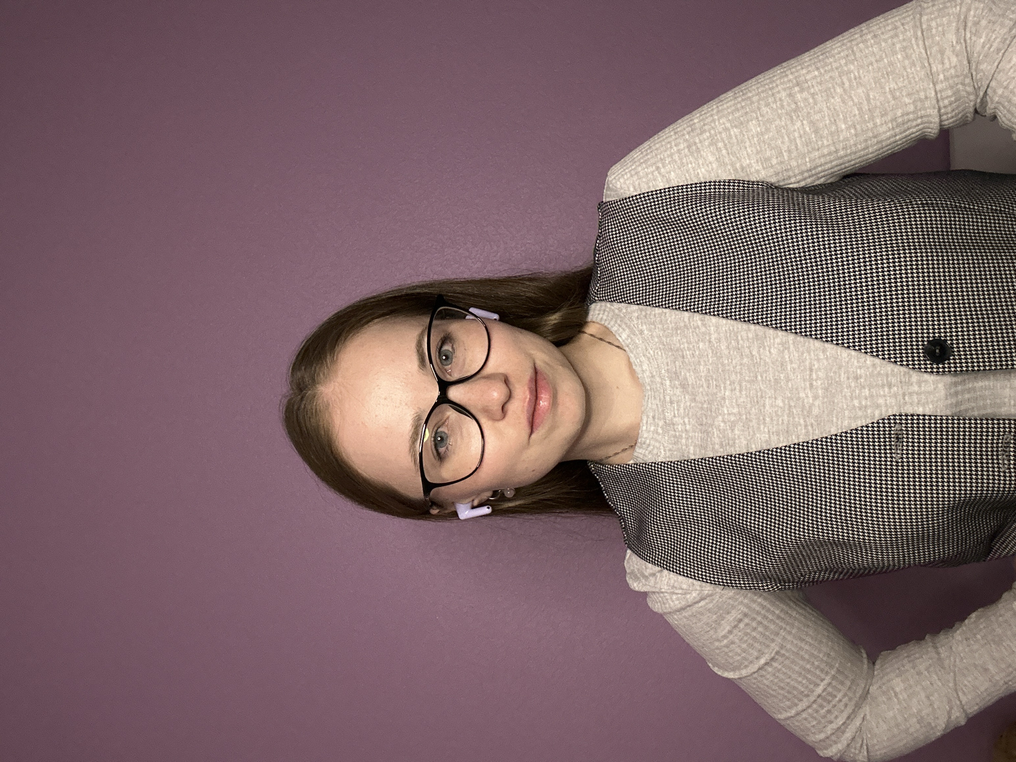 A young woman is depicted in a portrait-style image, facing forward with a gentle, neutral expression. She has light brown hair, parted on the side, and is wearing black-framed glasses that highlight her light-colored eyes. Her lips are slightly parted, revealing a natural lip color.
She is dressed in a light grey, long-sleeved top with a ribbed texture. Over this, she wears a sleeveless garment or vest featuring a distinct black and white houndstooth pattern. A single black button is visible on the front of the houndstooth piece, positioned on her left side (viewer's right). Light purple wireless earbuds are visible in both of her ears.
The background is a plain, solid muted purple wall, indicating an indoor setting. The lighting is soft and even, suggesting artificial light. There are no other discernible objects or details in the background. No specific time of day or weather conditions can be determined from the image. The context provided for the image is Saint Petersburg, Russia. There is no visible text in the media file.