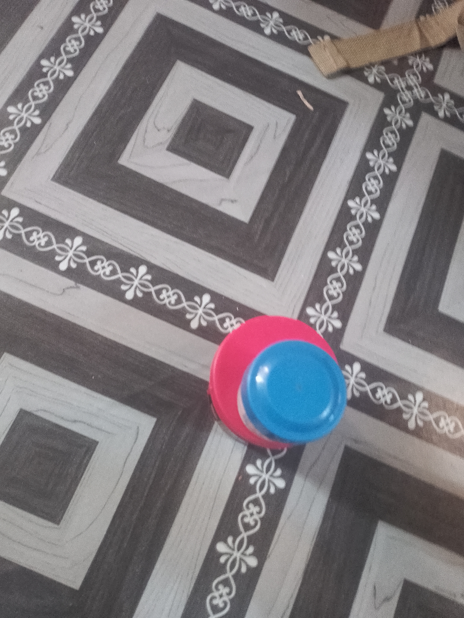 The image shows a close-up, overhead view of two plastic lids lying on a patterned floor. The floor has a geometric design with alternating squares of dark gray and light gray, outlined by a decorative white border. One lid is bright pink and is on the bottom, while a smaller blue lid is placed on top of it. The lighting suggests it is daytime, possibly indoors given the appearance of the floor which could be linoleum or tile. There are no people or animals visible in the frame. The overall scene is simple and focused on the objects and the floor's pattern. There is a strip of what appears to be burlap or a similar coarse fabric in the upper right corner of the image. No text is visible.