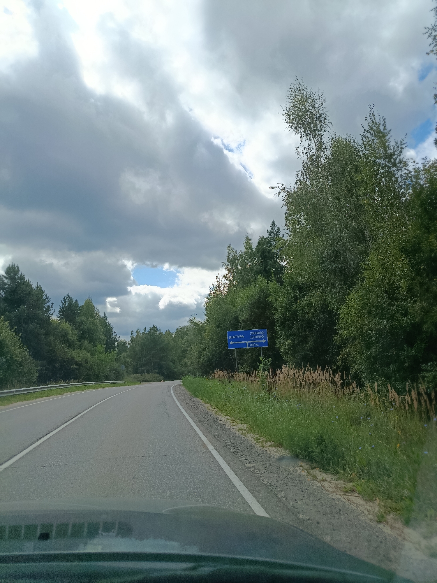 The image shows a road scene in Gribchikha, Russia, with a blue directional sign. The sign indicates "SHATURA" to the left with an arrow pointing left, and "LIKINO-DULEVO" with an arrow pointing right, followed by "350m". The road is paved and curves to the right, bordered by a metal guardrail on the left and a grassy shoulder with tall, dry grass and some scattered blue flowers on the right. A dense forest of green trees lines both sides of the road, with some taller, lighter-colored trees in the background. The sky is partly cloudy with dark gray clouds and patches of blue sky visible. The photo appears to be taken from a moving vehicle, as indicated by the reflection on the dashboard in the foreground. There are no people or animals visible in the image.