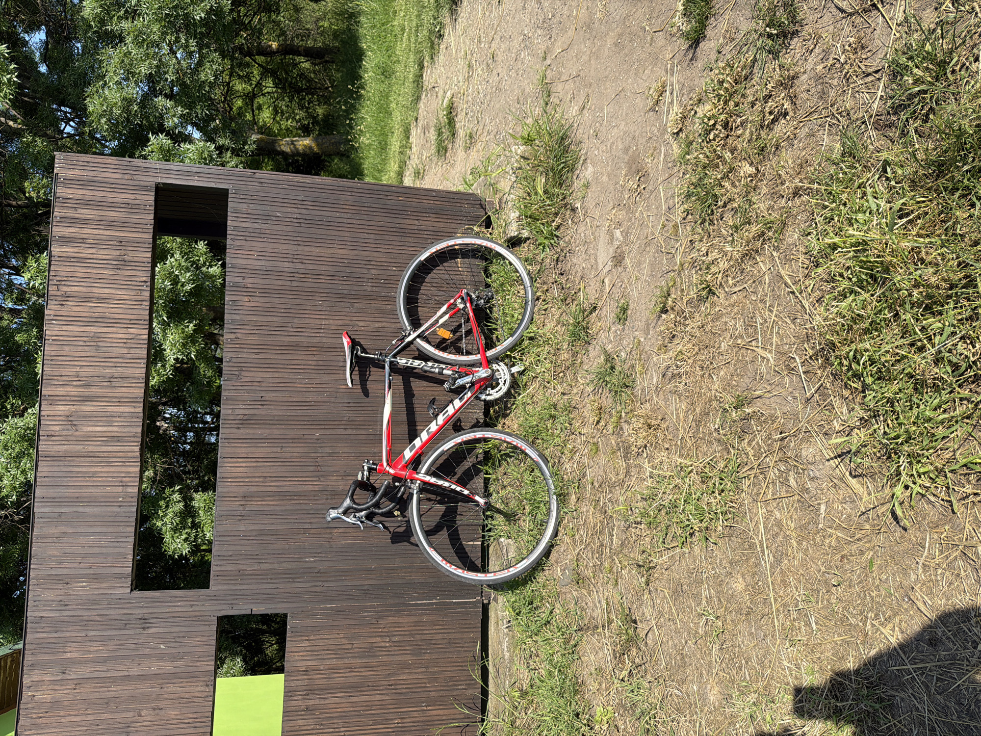 A red and white road bicycle is leaning against a brown wooden structure in a park-like setting. The structure has two rectangular openings, one above the other, and a section painted green at the bottom. The ground is a mix of grass and dirt, with some dry patches. Trees are visible in the background. It is daytime with bright sunlight. The location is Burgas, Bulgaria.