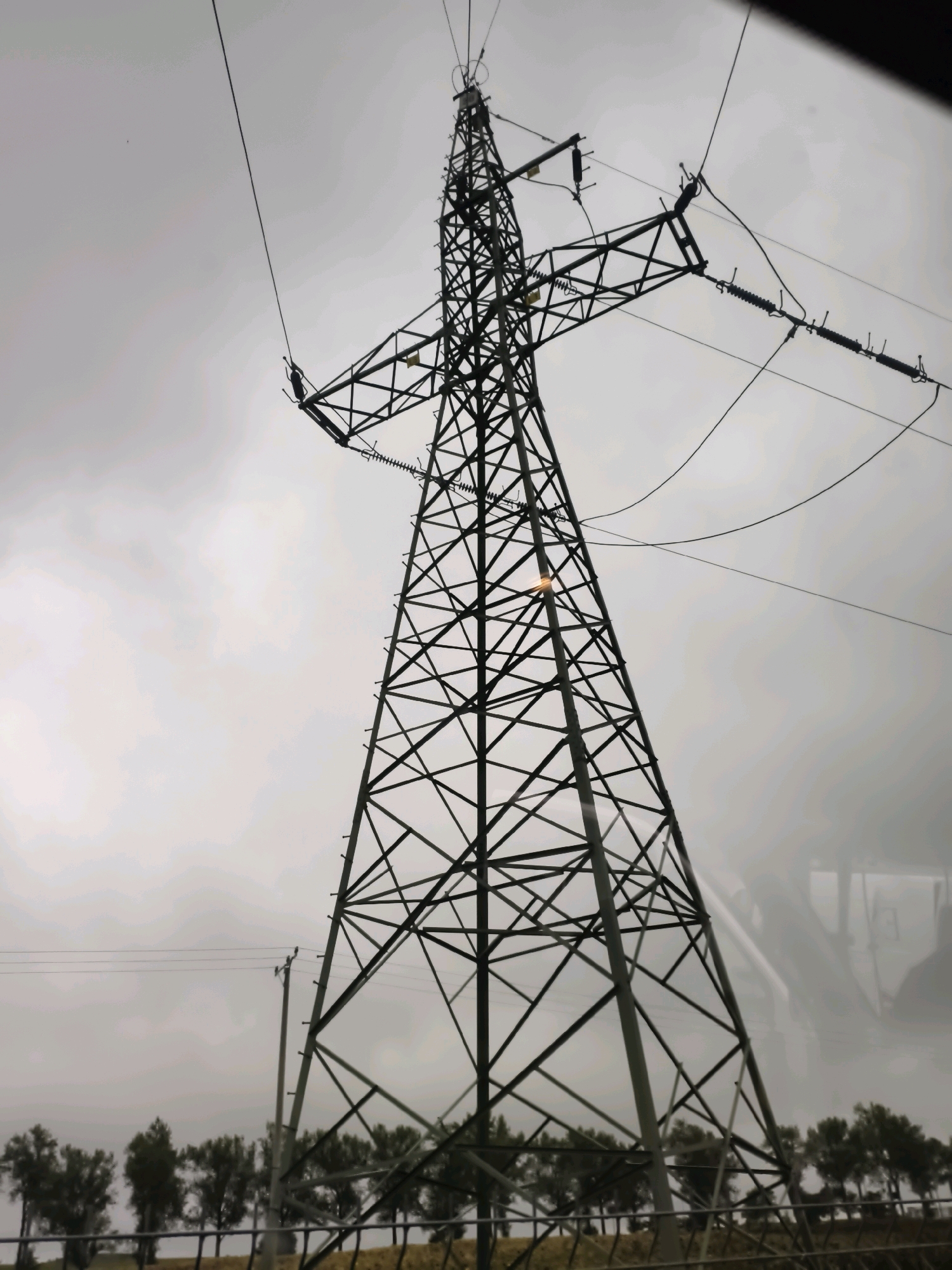 This image depicts a high-voltage electricity pylon against a cloudy sky. The pylon is constructed of dark metal lattice work. Several power lines extend from the top of the pylon. In the background, a row of trees is visible, with a fence in front of them. A smaller utility pole is situated to the left of the pylon. The overall scene suggests an industrial or rural setting, possibly on the outskirts of a city.