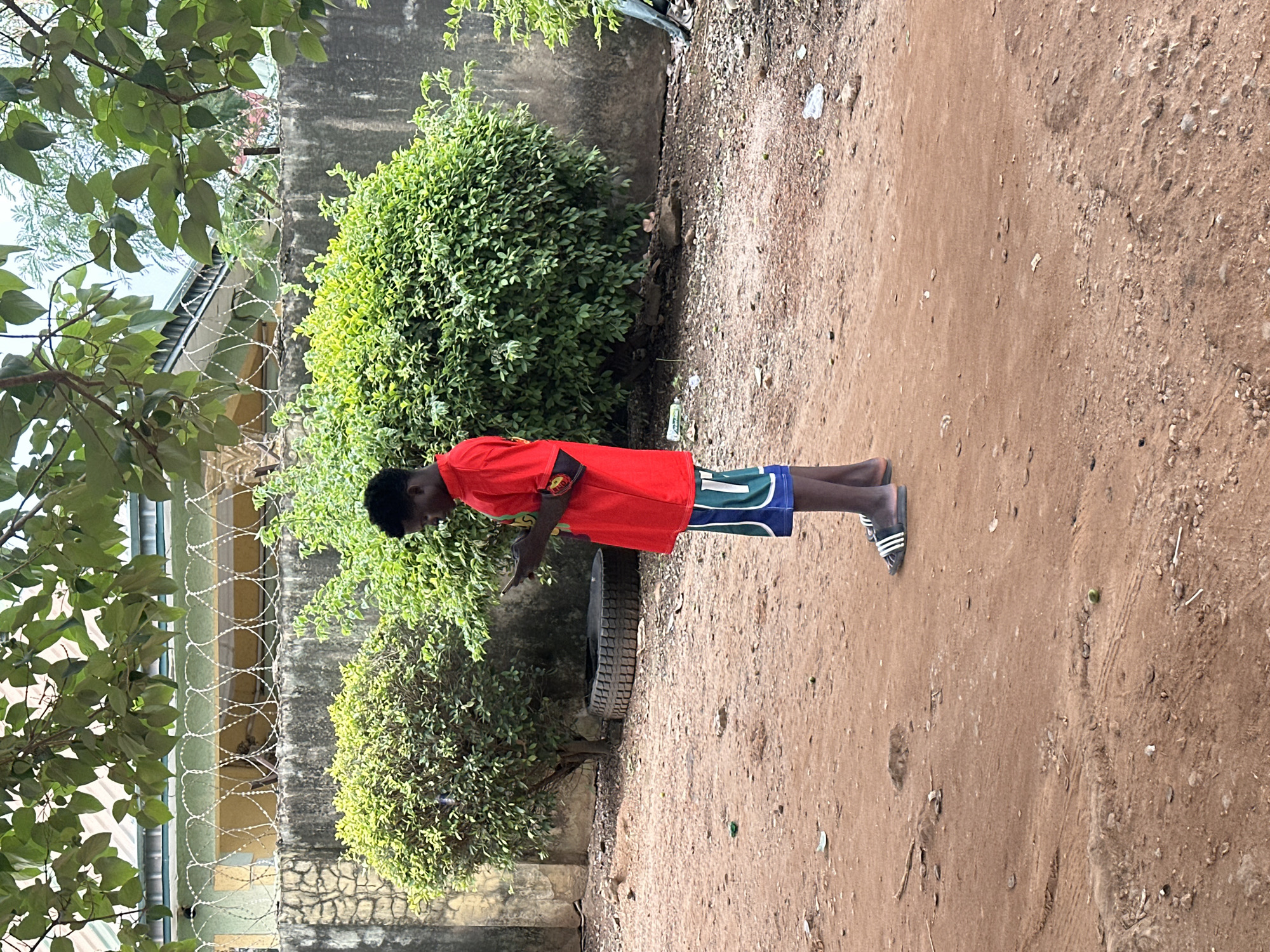 A young Black male is standing in a dusty, outdoor area, looking at a mobile phone. He is wearing a bright red t-shirt and blue and green patterned shorts. On his feet, he has black and white striped slides. Behind him, there is a concrete wall topped with barbed wire, and beyond that, a building with a light green facade and a metal roof. Lush green bushes are visible in front of the wall. To the left of the boy, a tire is lying on its side. The ground is covered in reddish-brown dirt with scattered debris, including leaves and small rocks. The overall scene suggests an outdoor, possibly residential or semi-developed area in Abuja, Nigeria.