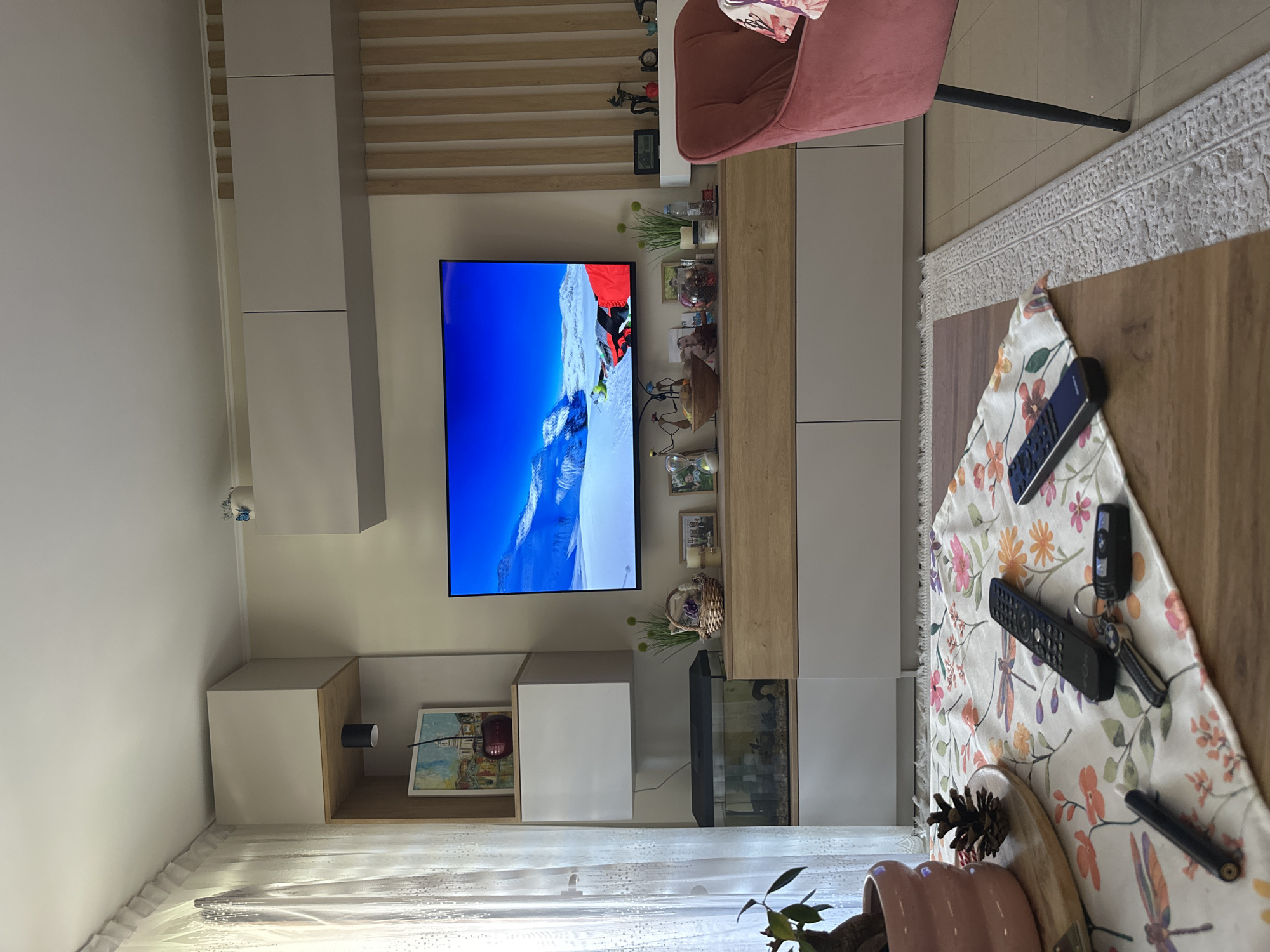 The image presents an interior scene, possibly a living room, featuring a contemporary design with light-colored furniture. A large television displays a mountainous landscape with a skier, mounted on the wall. To the side of the television, there's a decorative shelving unit displaying various ornaments and small plants. Below the TV, a painting is placed next to a fish tank. A floral-patterned runner lies on a wooden coffee table, with multiple remote controls, a key, and a pine cone on it. A pink chair is partially visible in the upper right corner of the frame. Light permeates through sheer curtains in the bottom left. The precise location within Plovdiv, Bulgaria, cannot be determined from this image alone.