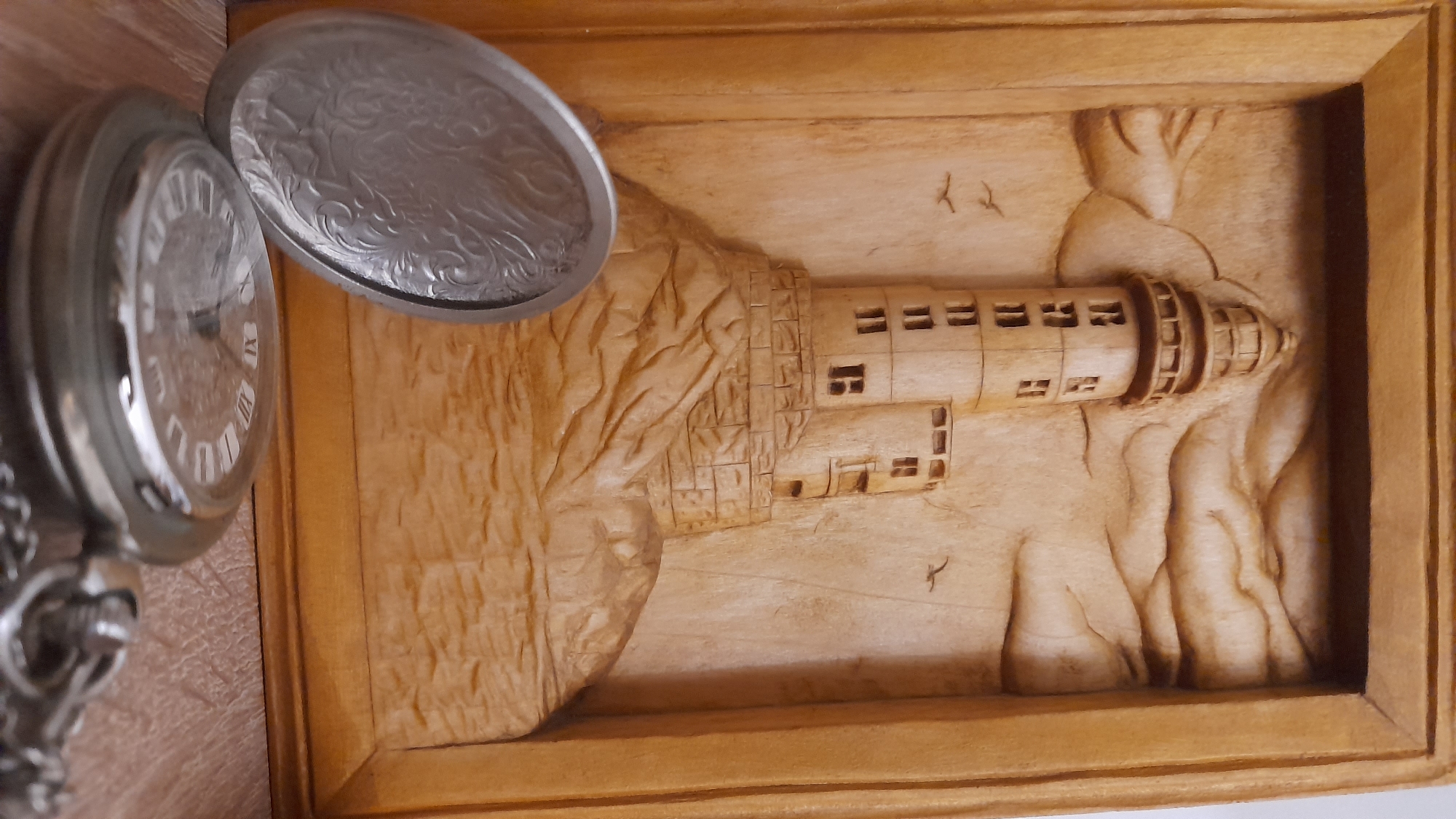 The image shows a close-up of a silver pocket watch resting on a wooden carving of a lighthouse scene. The pocket watch is partially open, revealing a clock face with Roman numerals. The wooden carving depicts a lighthouse built on a rocky cliff face. Two small birds appear to be etched into the background. There is no indication this is in Saint Petersburg.