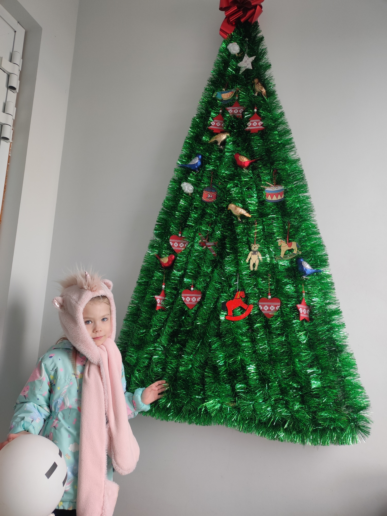 A young girl stands next to a green Christmas tree. The girl wears a pink unicorn hat and scarf, and a blue jacket with clouds on it. She holds a white balloon with a drawn-on face. The Christmas tree is decorated with red and gold ornaments in shapes of birds, hearts, stars, and drums. The tree is topped with a large red bow. The scene appears to be inside a room with white walls. The location is Votkinsk, Russia.